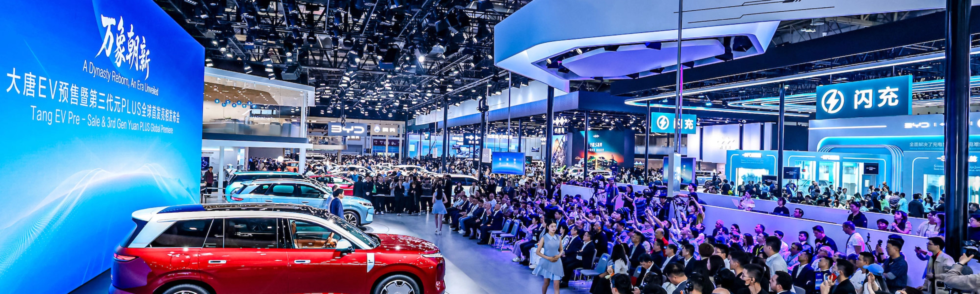 Red car on display at a crowded auto show with large screen.