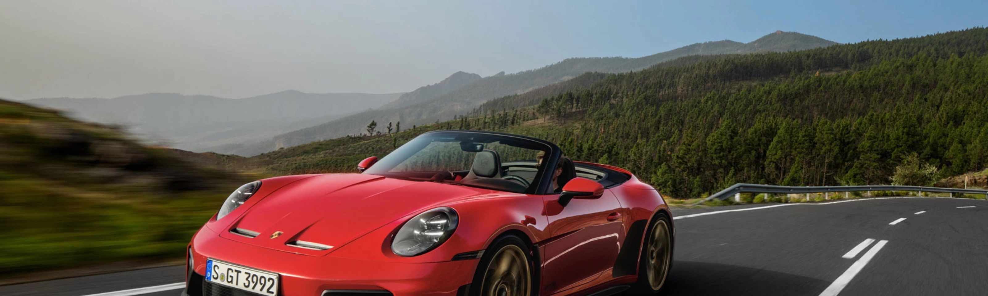 Red Porsche convertible driving on a winding road through green hills.