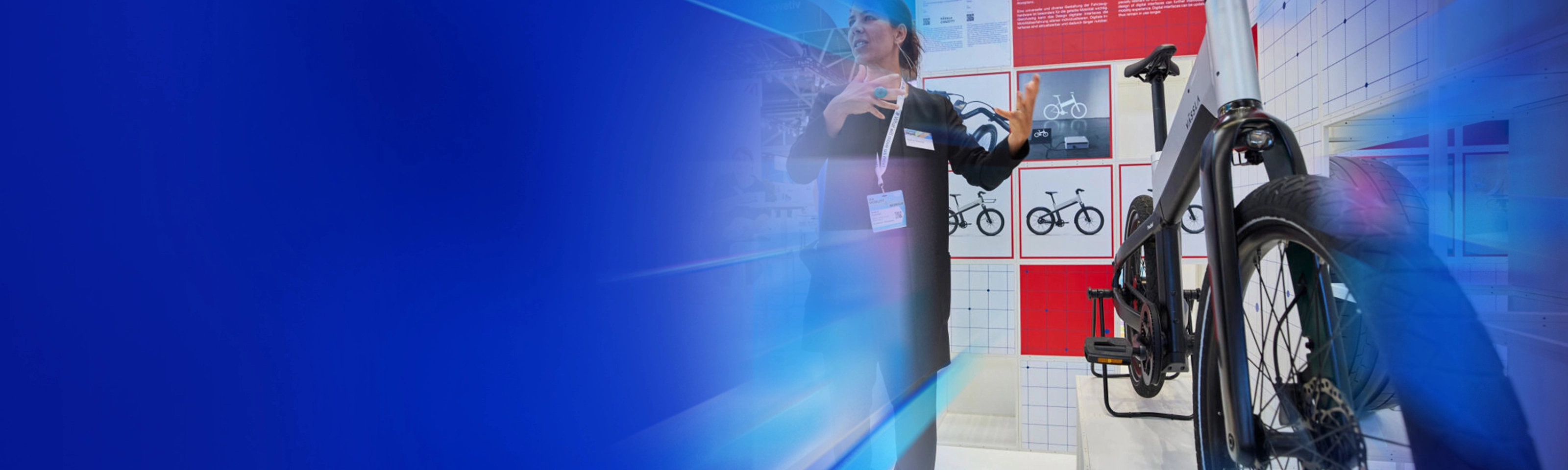 Eine Frau präsentiert ein modernes Fahrrad an einem Messestand mit Designpostern im Hintergrund.