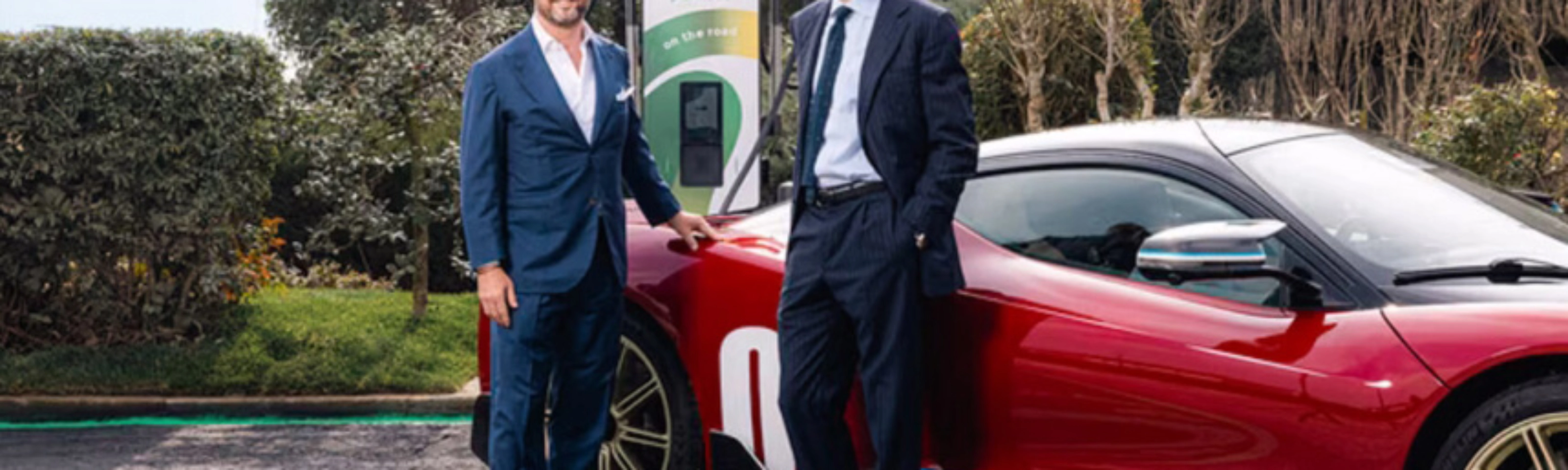Two men in suits stand beside a red electric sports car at a charging station.