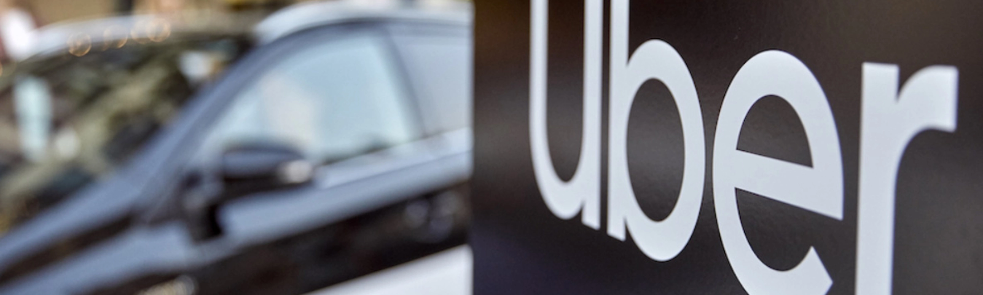Uber-Logo auf einem Schild, dahinter unscharf ein Auto.