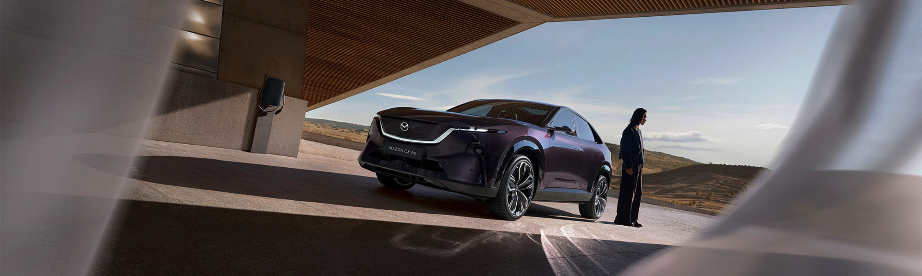 A person stands near a dark Mazda SUV under a modern building with open landscape behind.