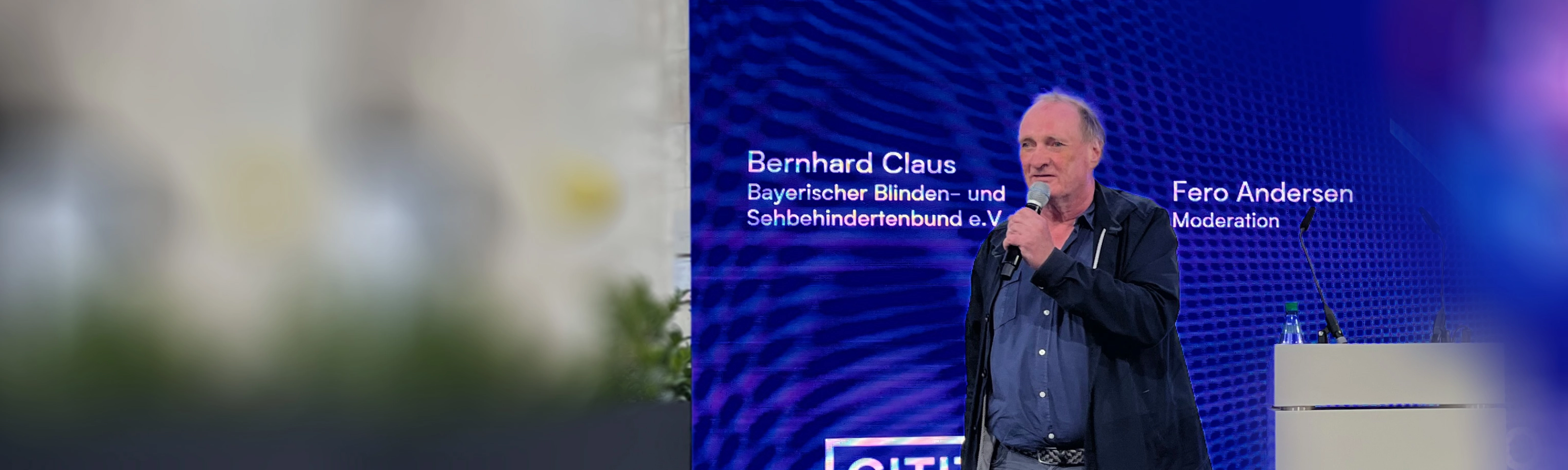 Ein Redner steht auf einer Bühne vor einem Bildschirm zur Mobilität für sehbehinderte Menschen.
