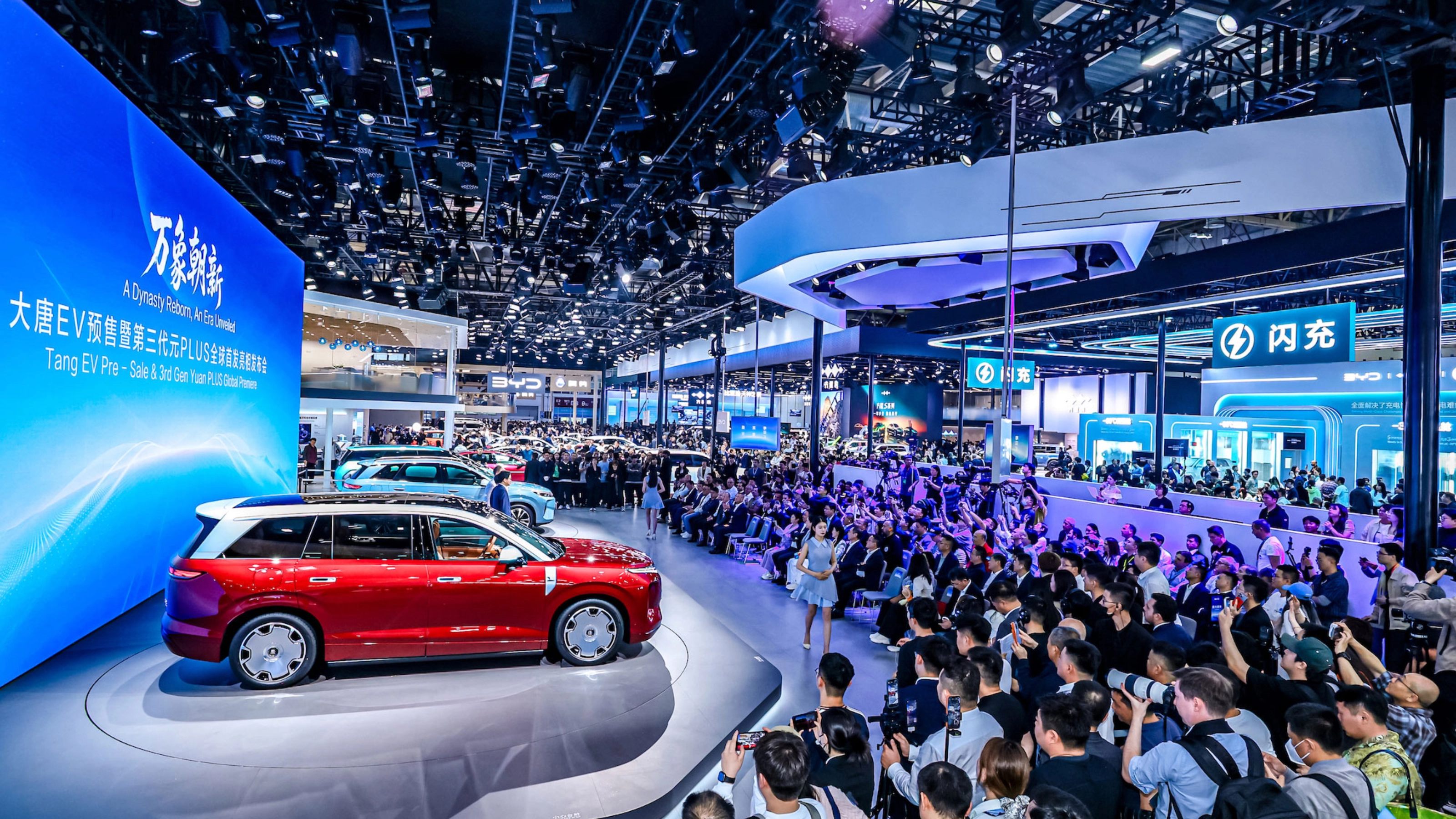 Red car on display at a crowded auto show with large screen.