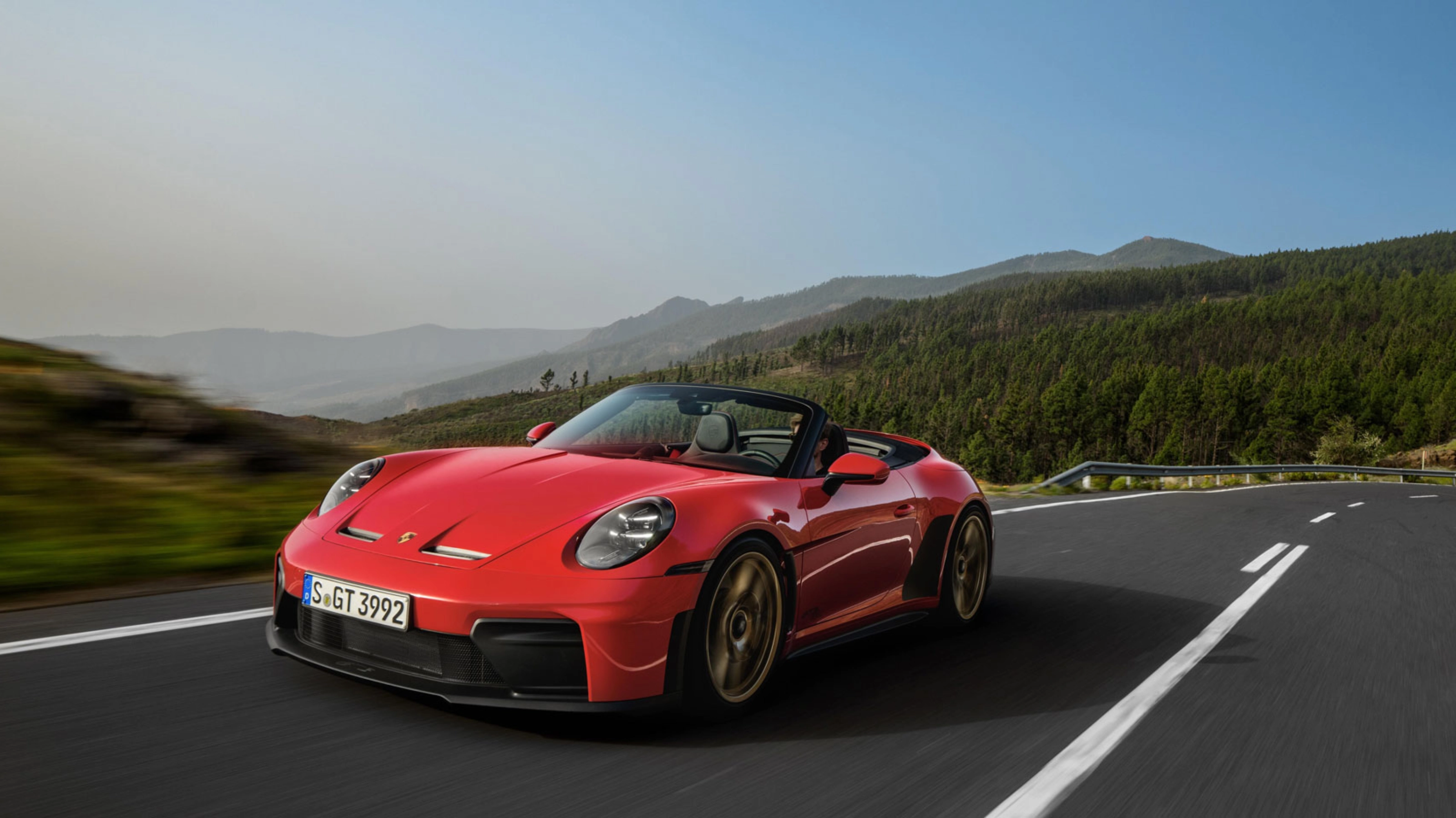 Red Porsche convertible driving on a winding road through green hills.