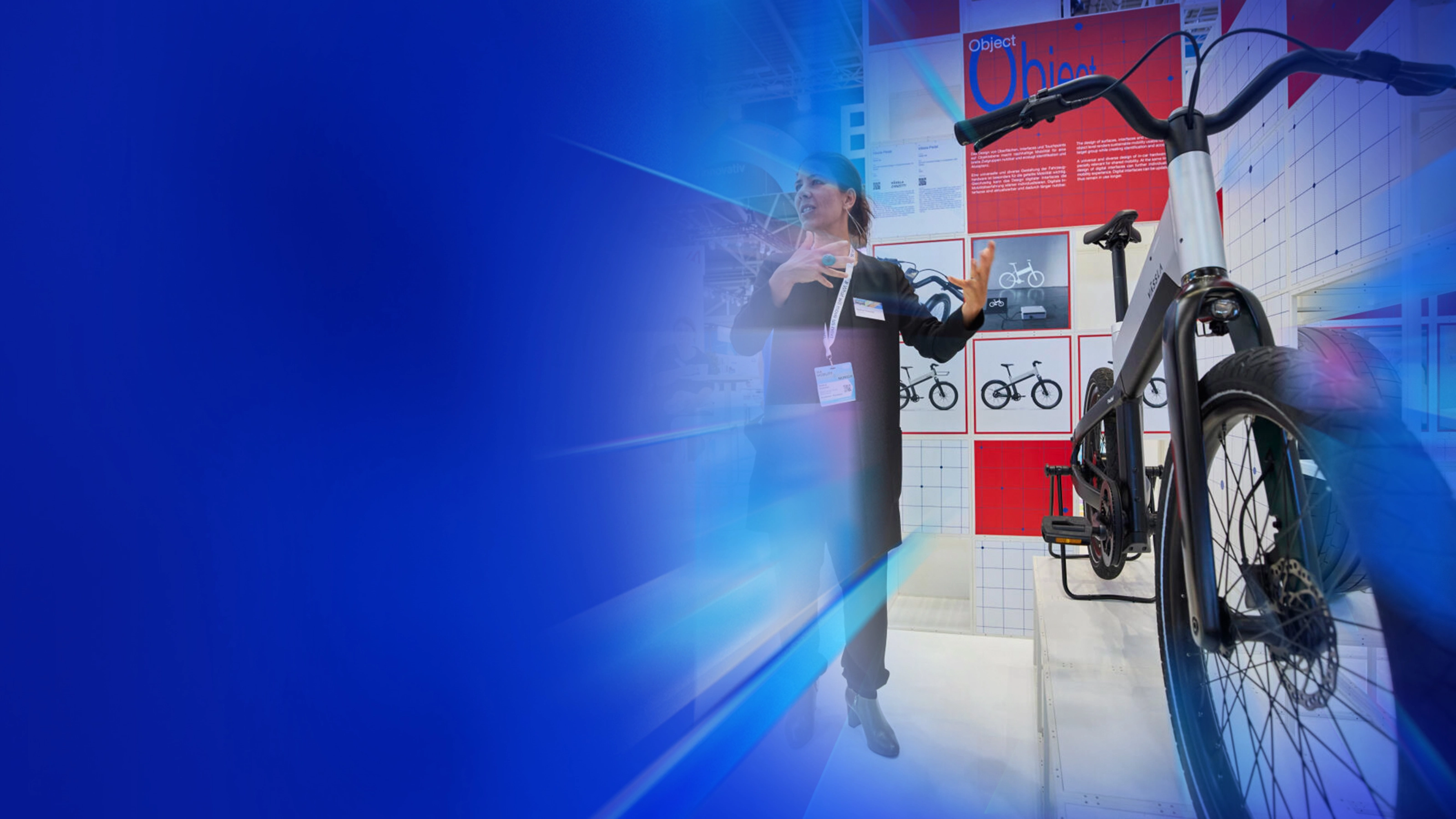 Eine Frau präsentiert ein modernes Fahrrad an einem Messestand mit Designpostern im Hintergrund.