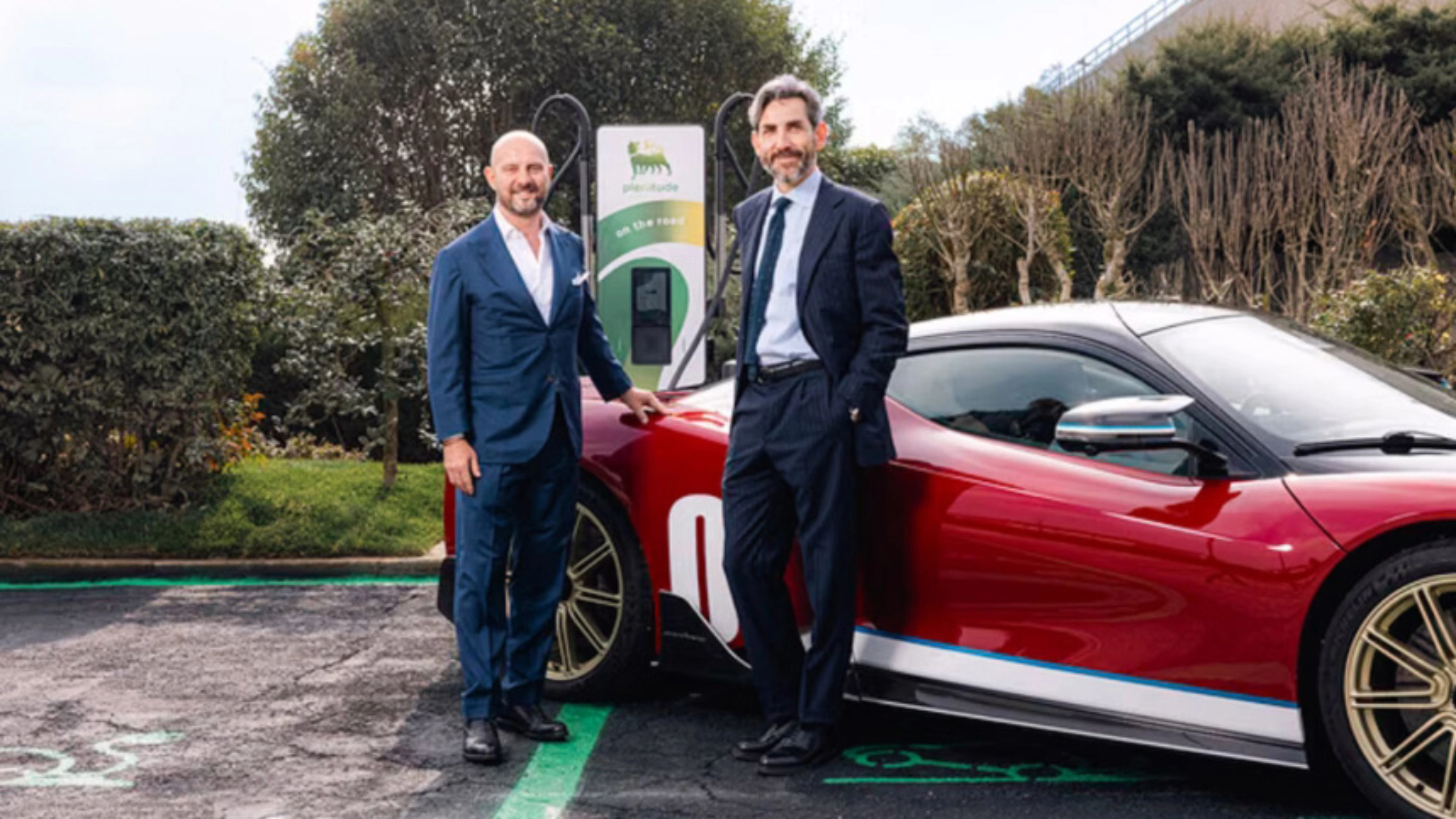 Two men in suits stand beside a red electric sports car at a charging station.
