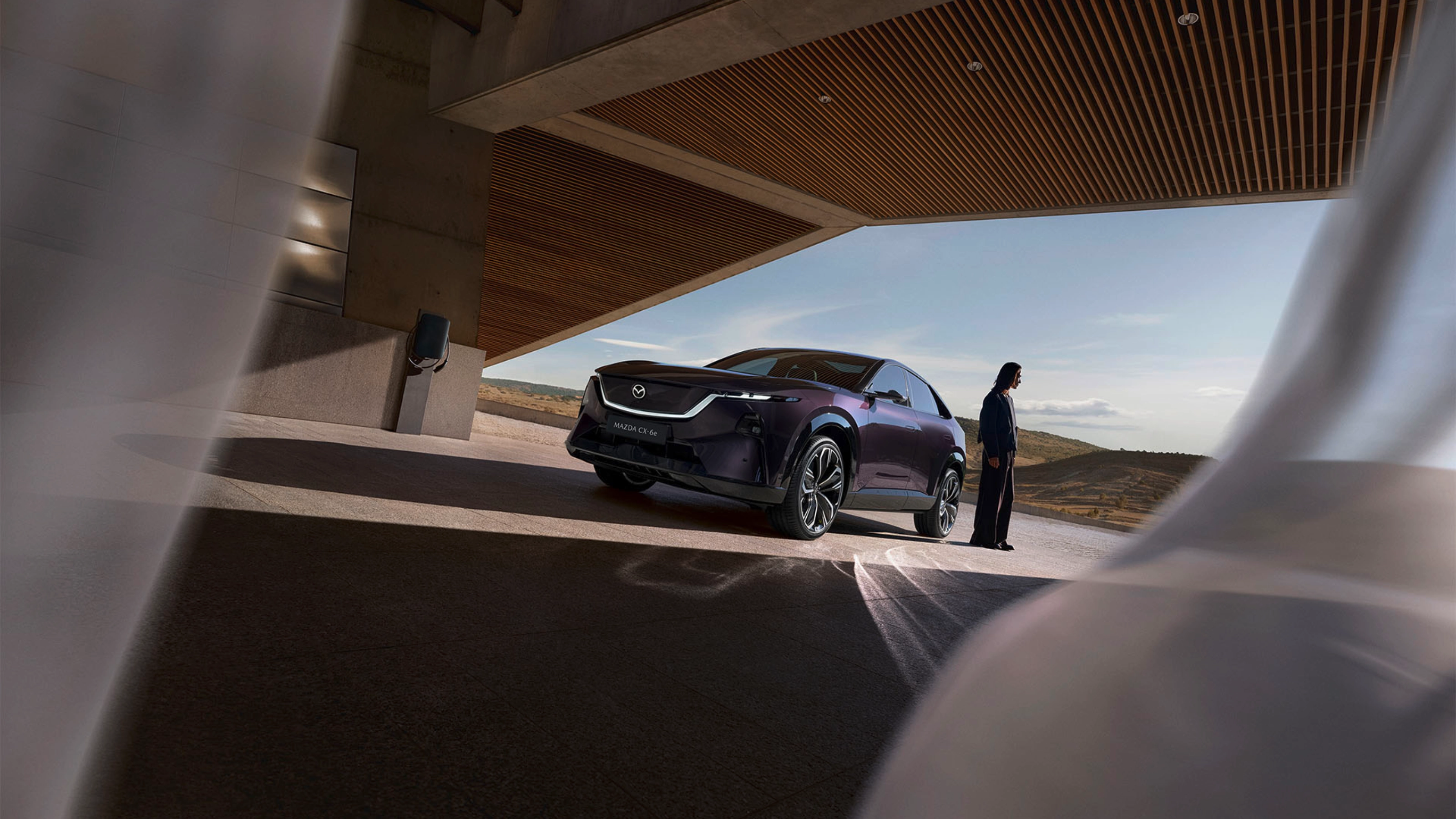 A person stands near a dark Mazda SUV under a modern building with open landscape behind.