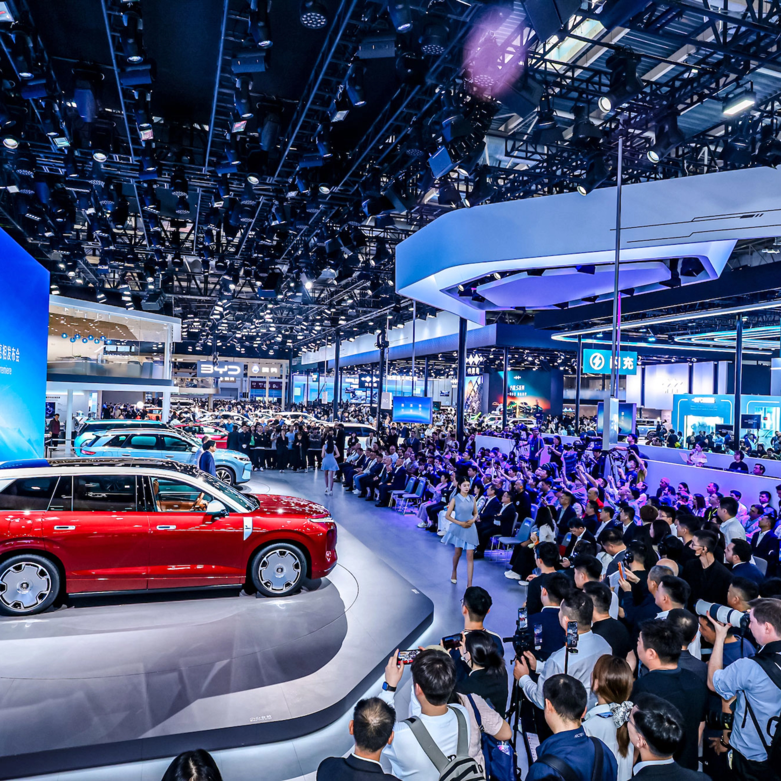 Red car on display at a crowded auto show with large screen.