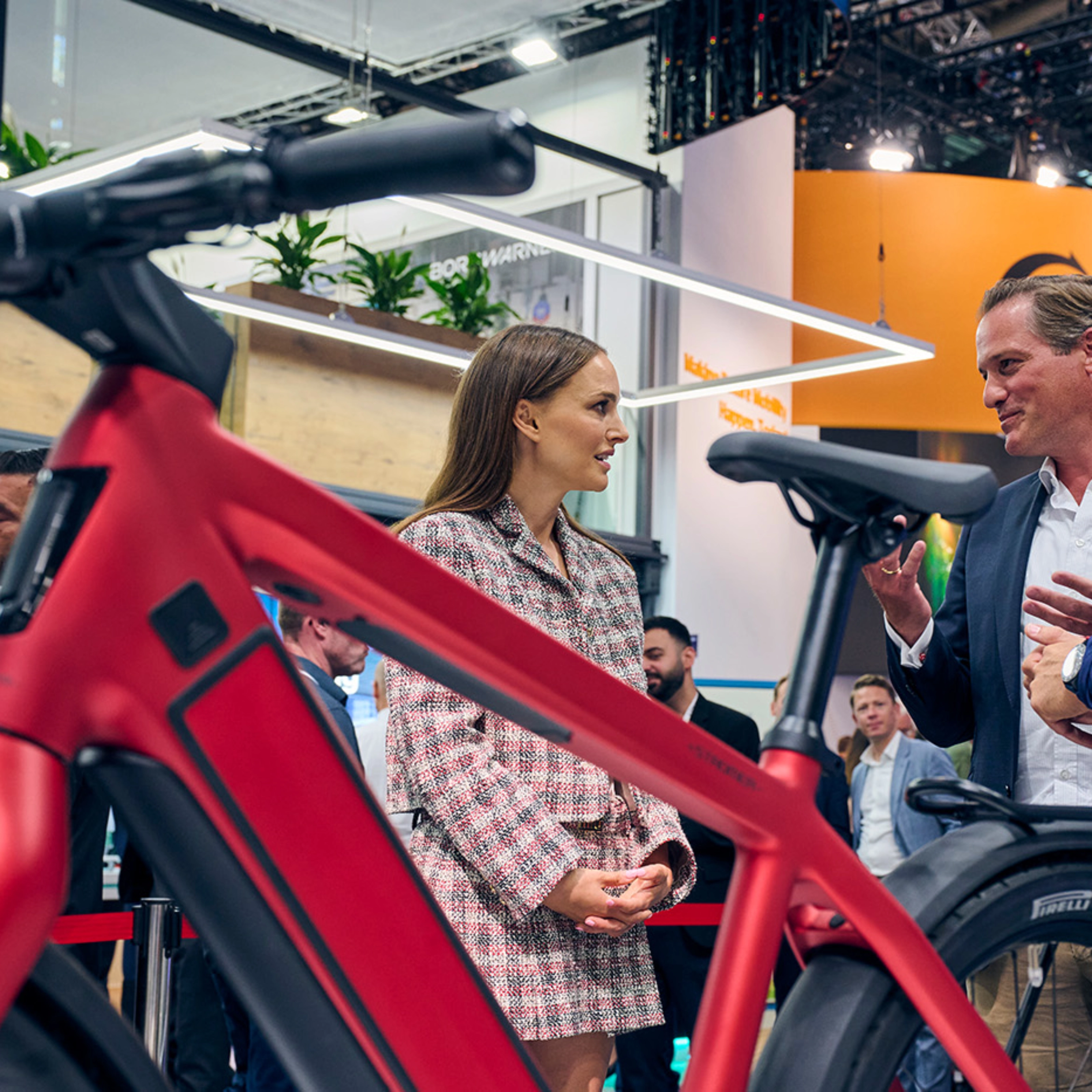 Natalie Portman in conversation with exhibitors at IAA Mobility 2023, standing near a red bicycle in a trade fair setting.