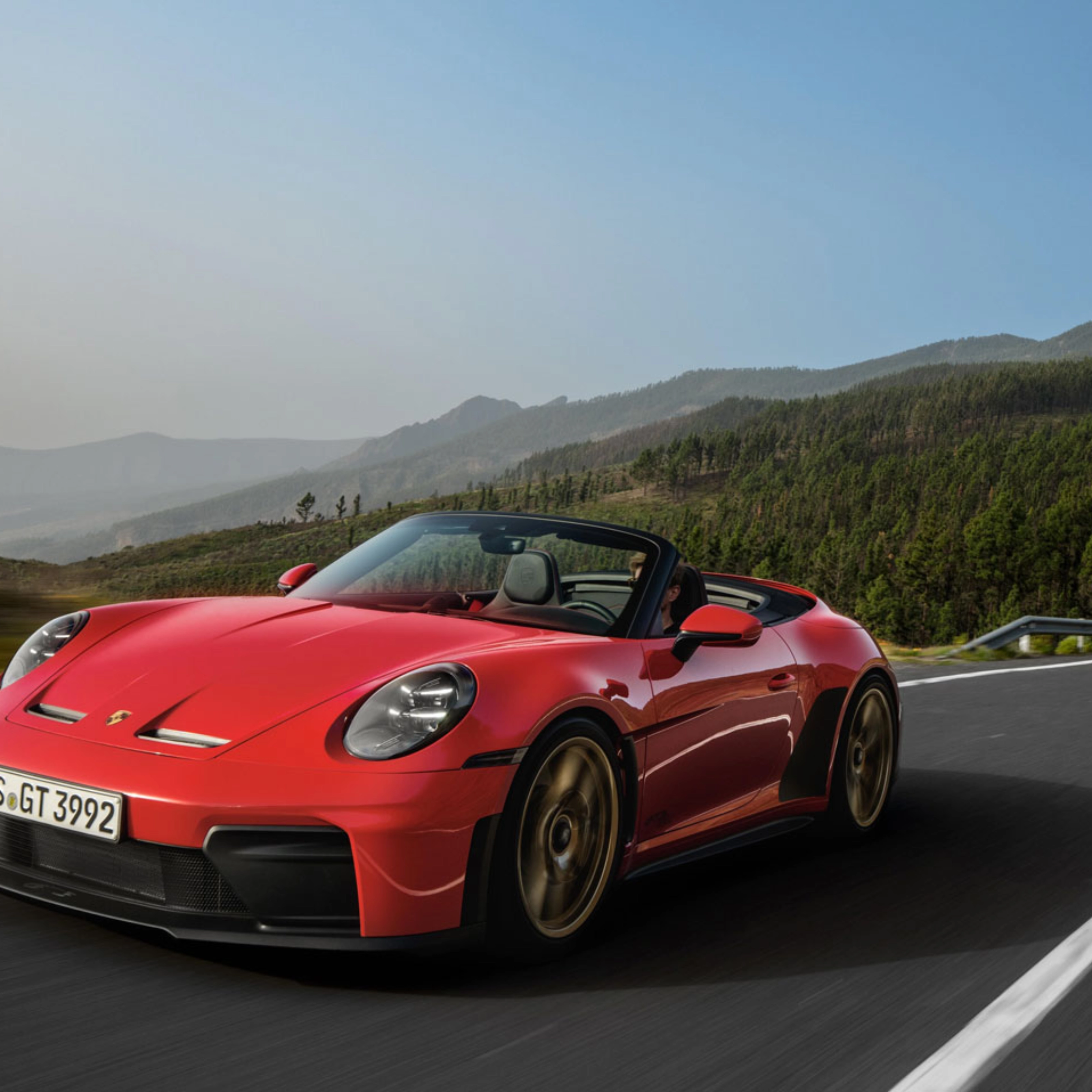 Red Porsche convertible driving on a winding road through green hills.
