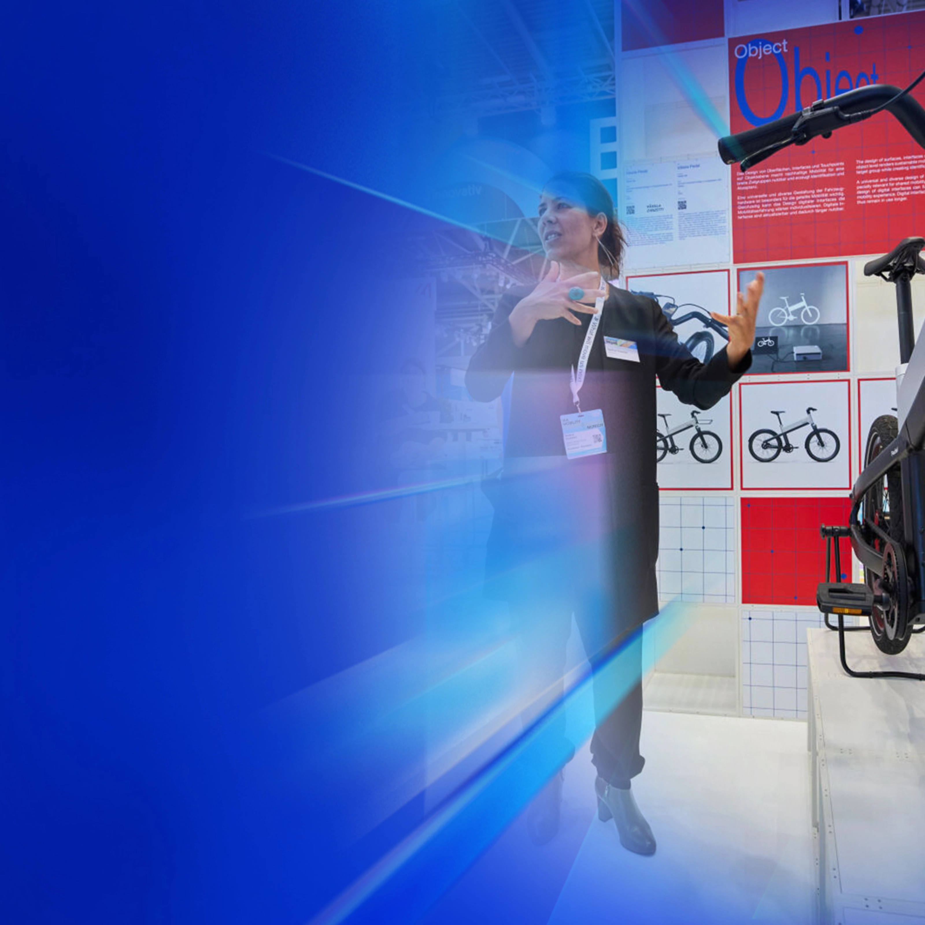 Eine Frau präsentiert ein modernes Fahrrad an einem Messestand mit Designpostern im Hintergrund.
