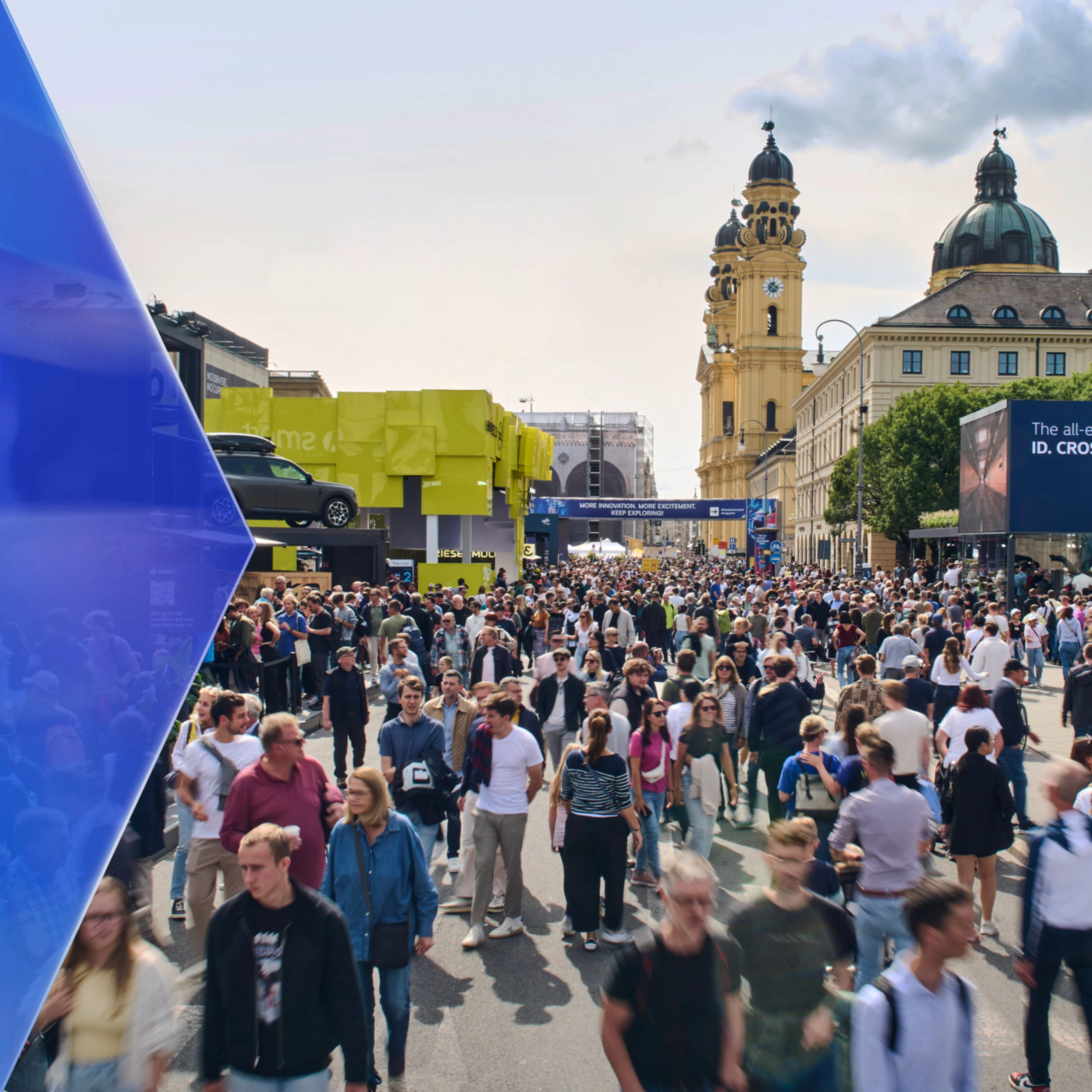 Große Menschenmenge auf einer Mobilitätsmesse im Freien mit Ausstellungsständen und Altbauten.