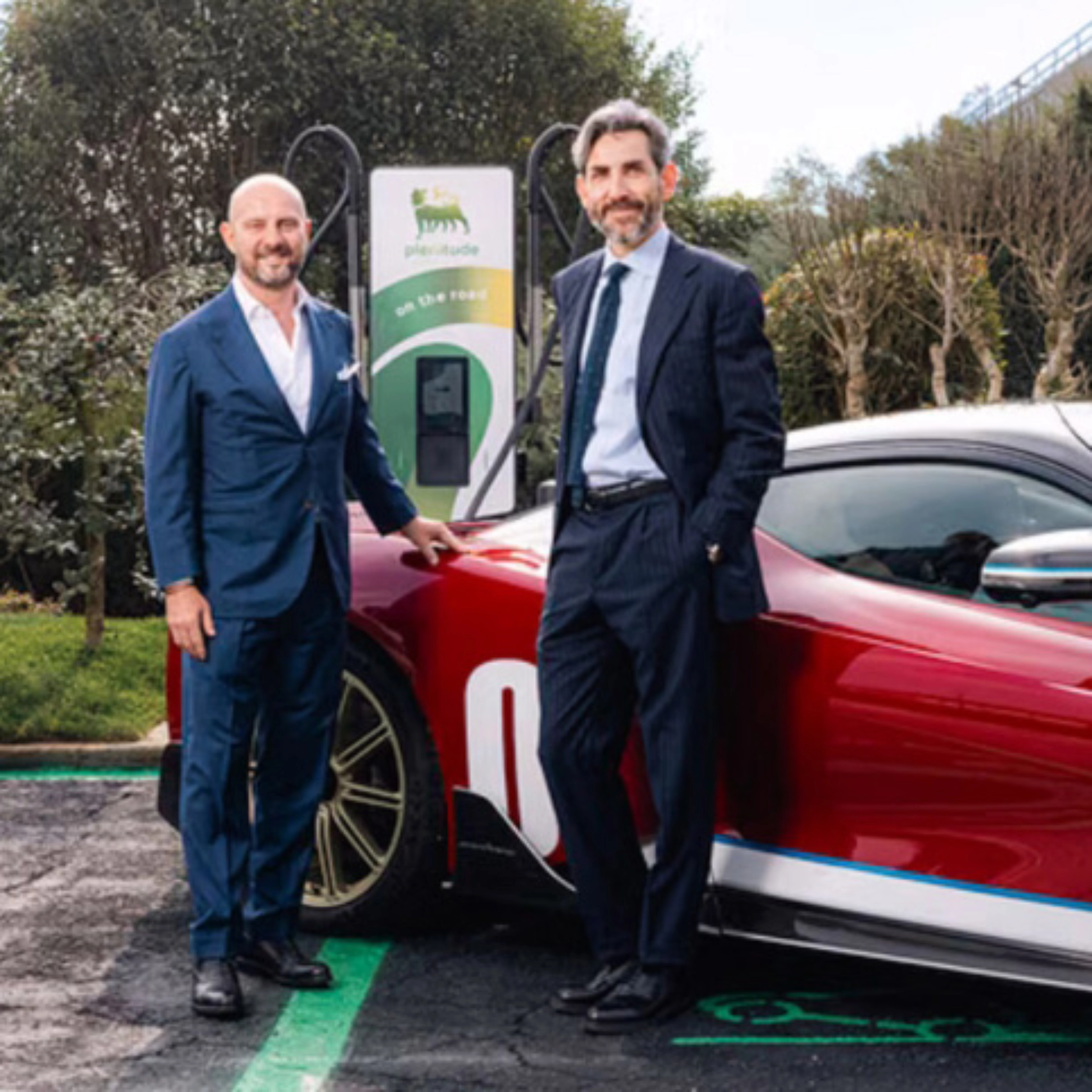 Two men in suits stand beside a red electric sports car at a charging station.