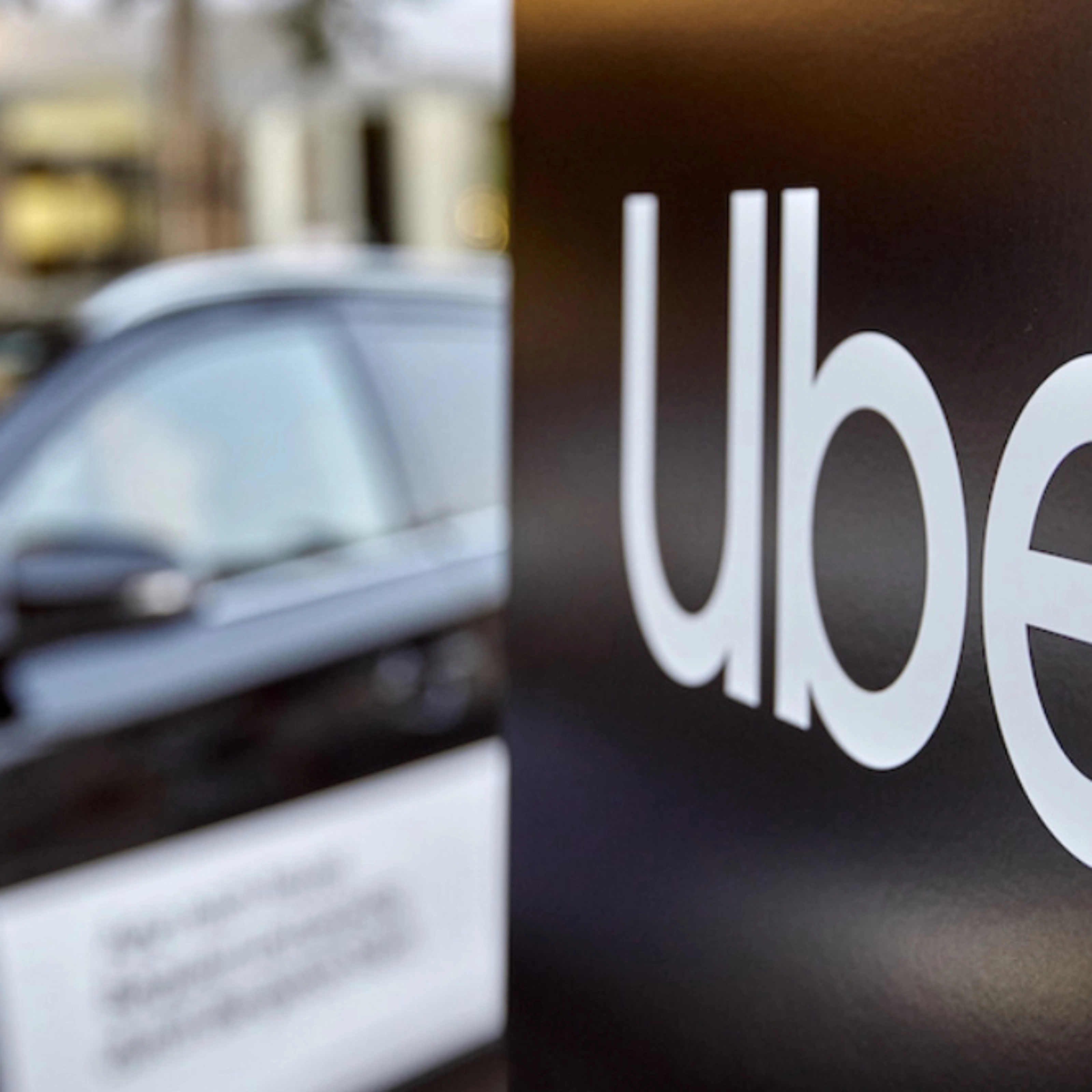 Uber-Logo auf einem Schild, dahinter unscharf ein Auto.