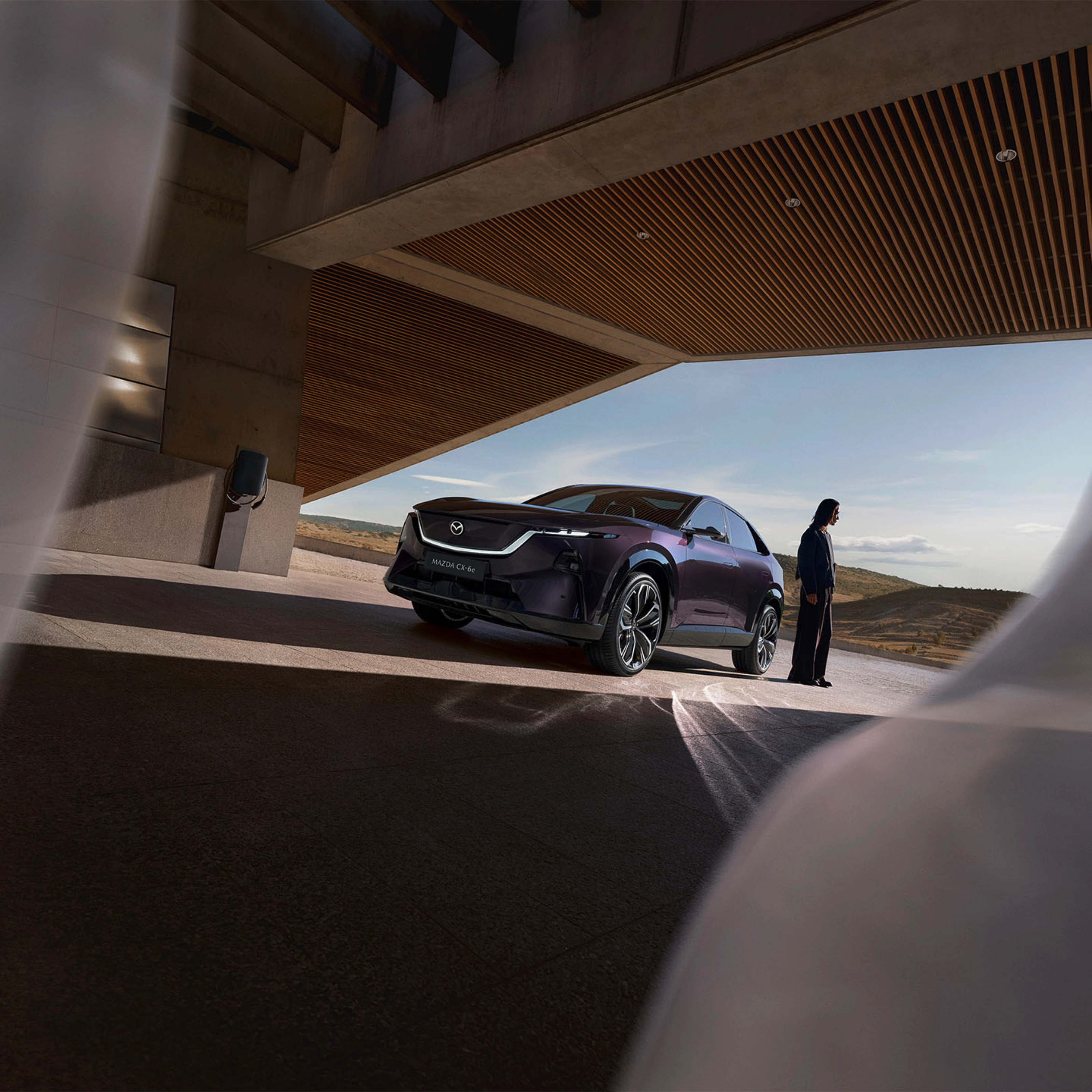 A person stands near a dark Mazda SUV under a modern building with open landscape behind.