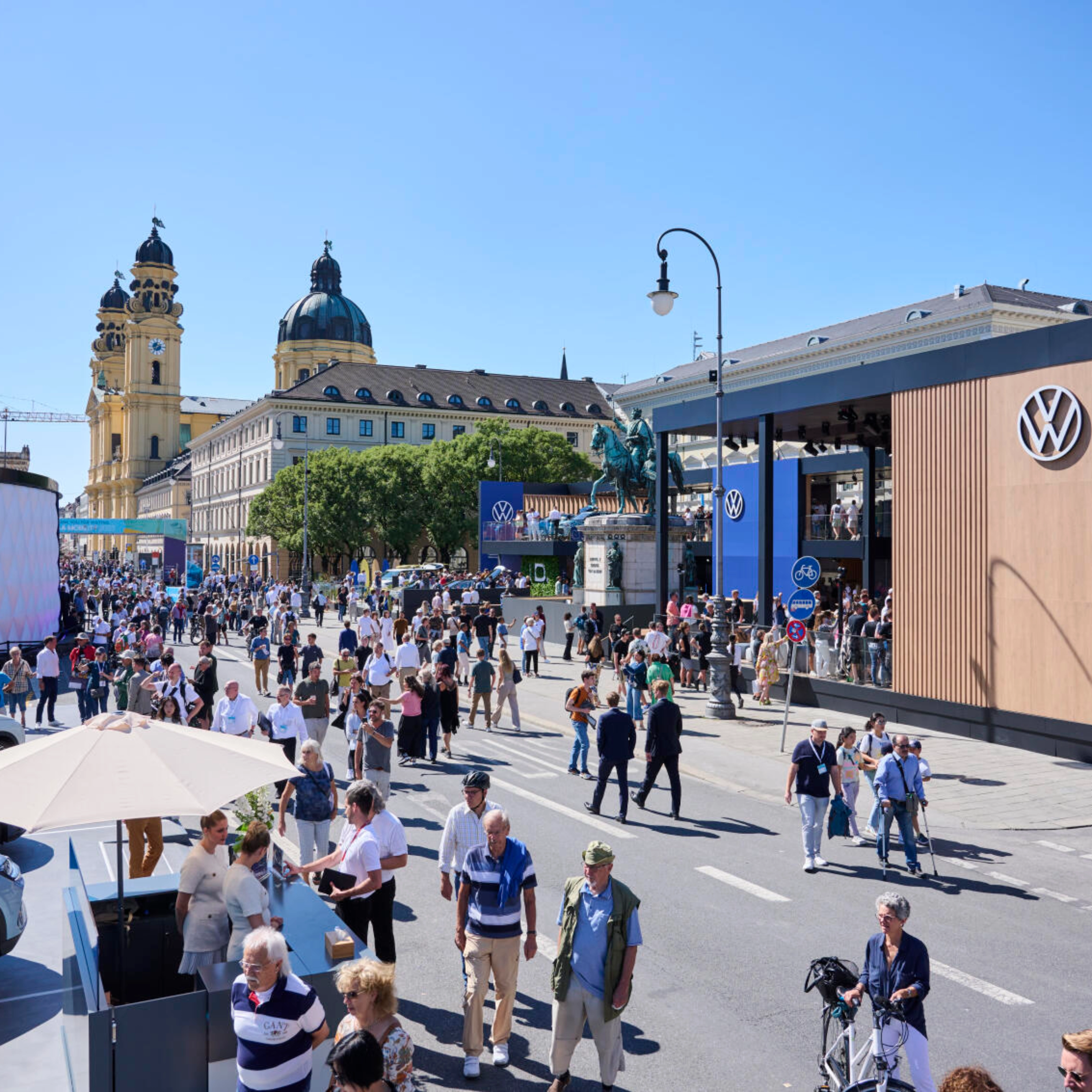 Menschen besuchen eine Outdoor-Mobilitätsmesse mit Ständen und historischen Gebäuden.