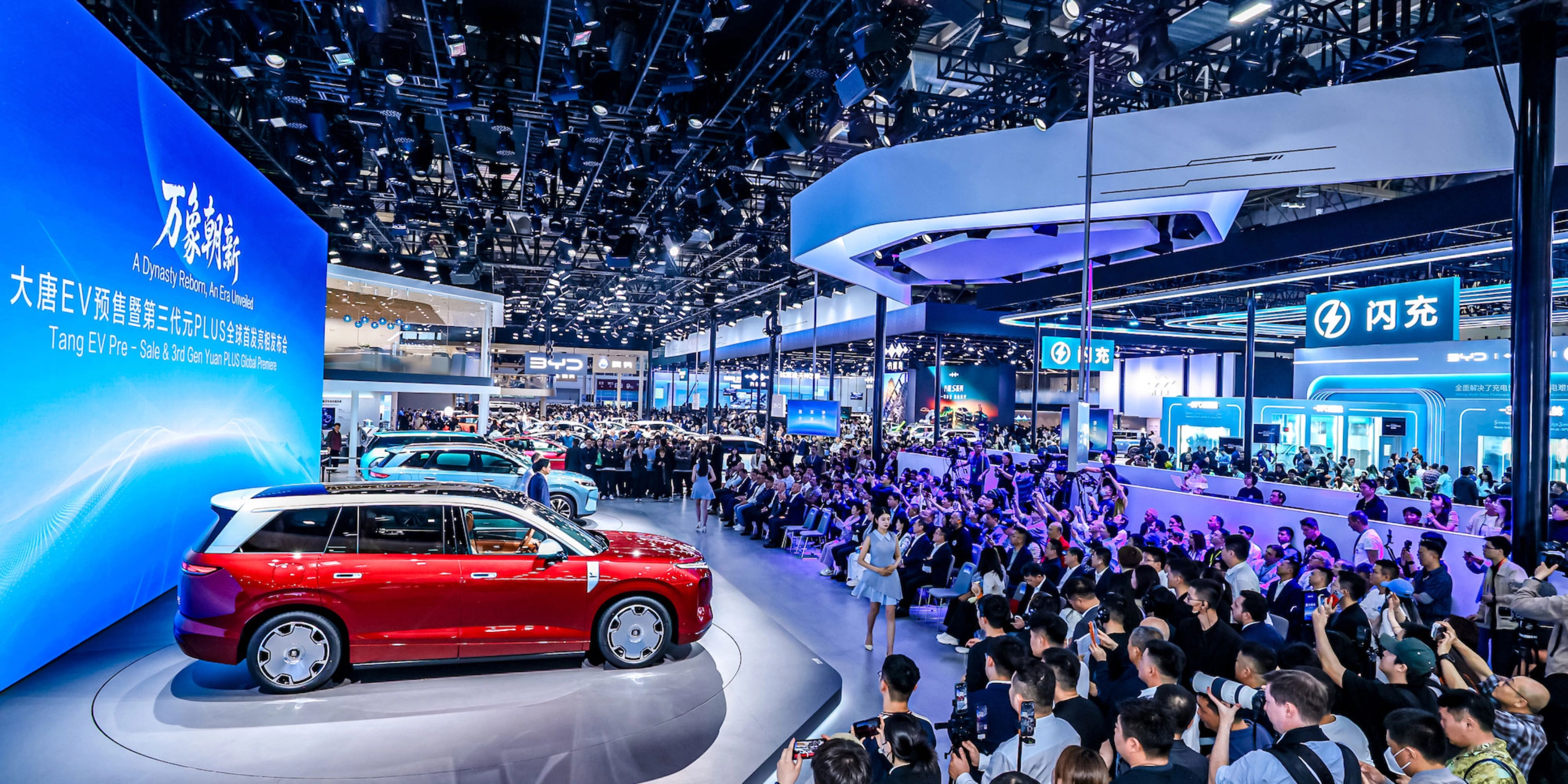 Red car on display at a crowded auto show with large screen.