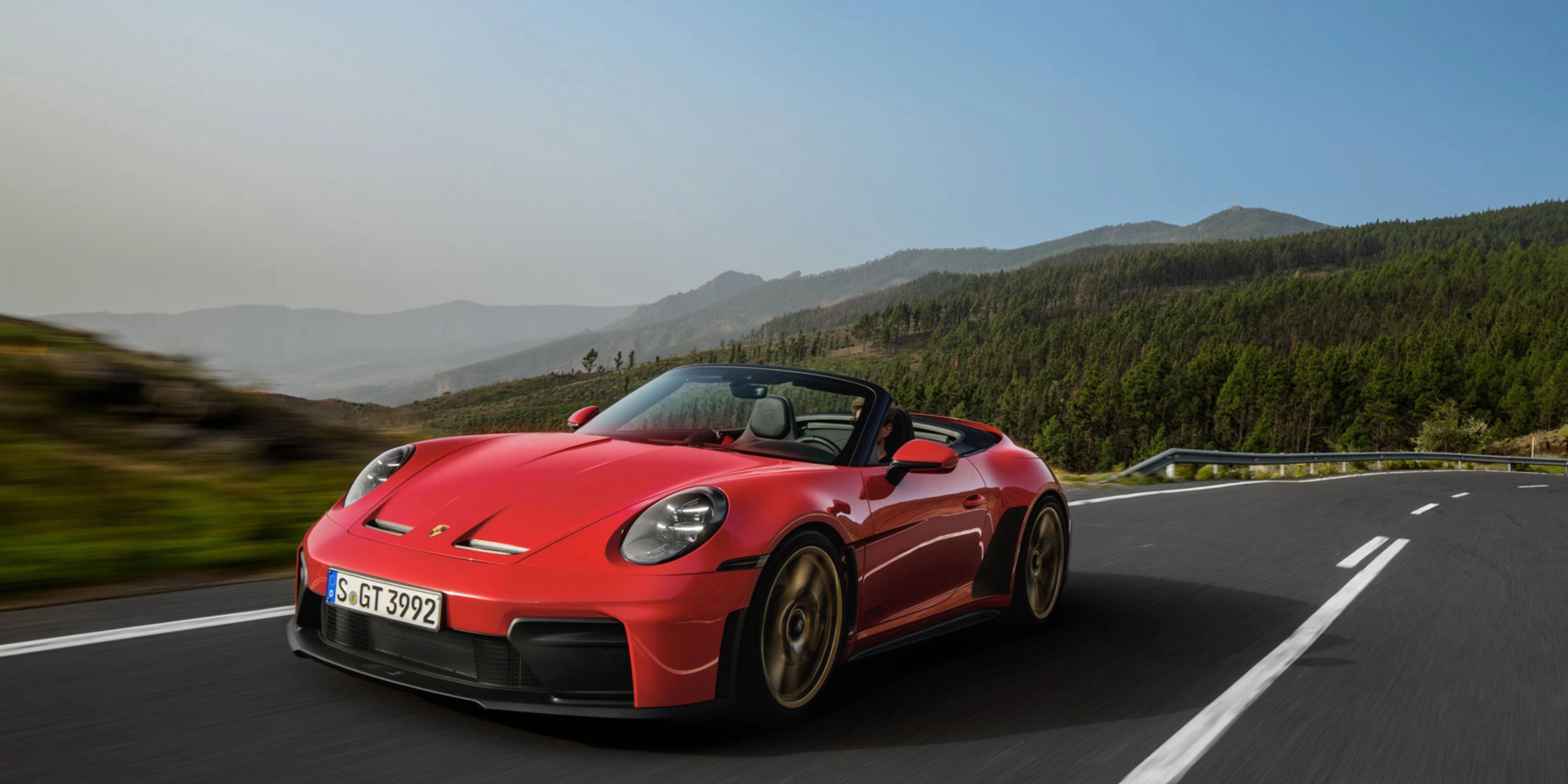 Red Porsche convertible driving on a winding road through green hills.