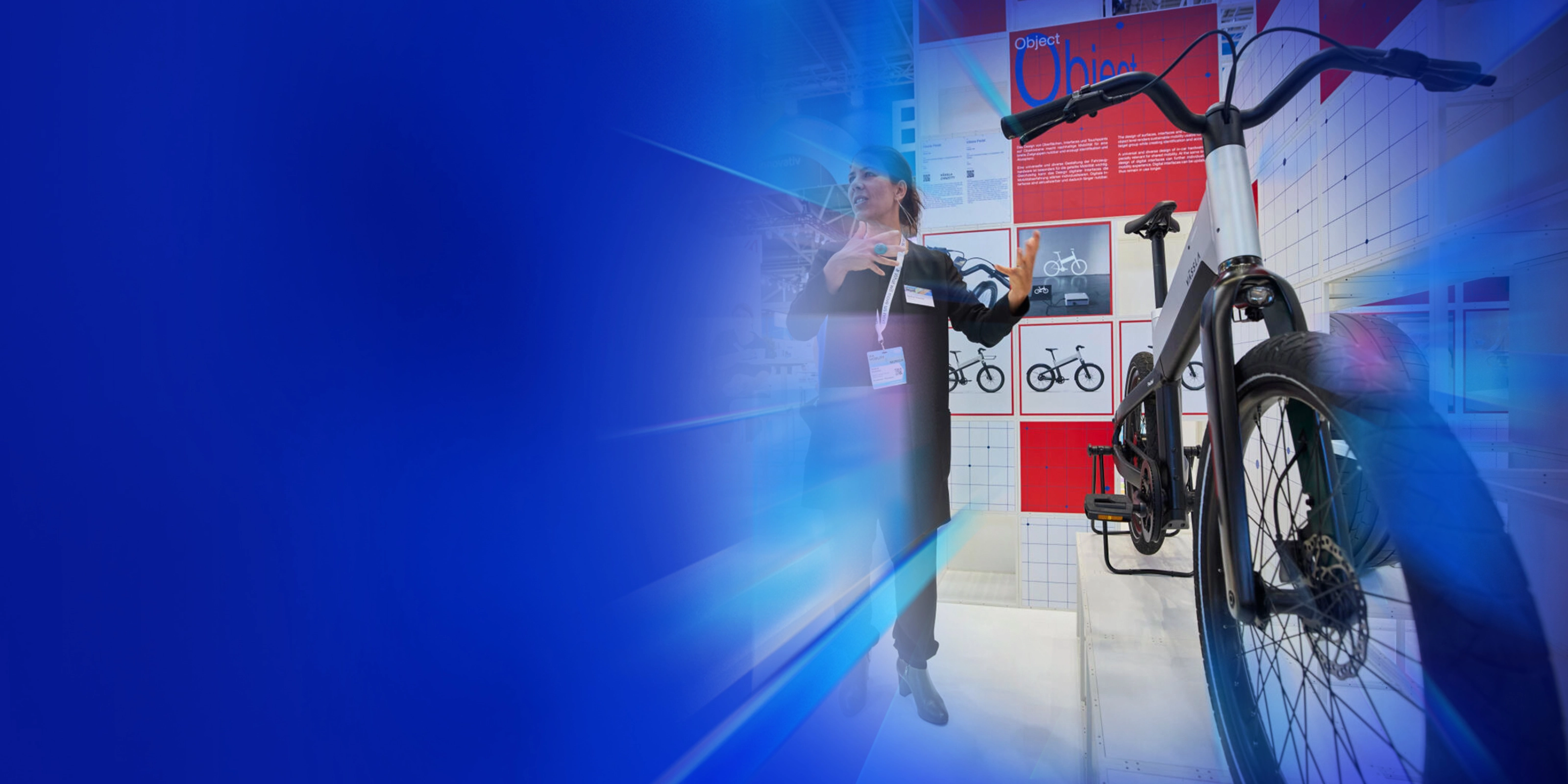 Eine Frau präsentiert ein modernes Fahrrad an einem Messestand mit Designpostern im Hintergrund.