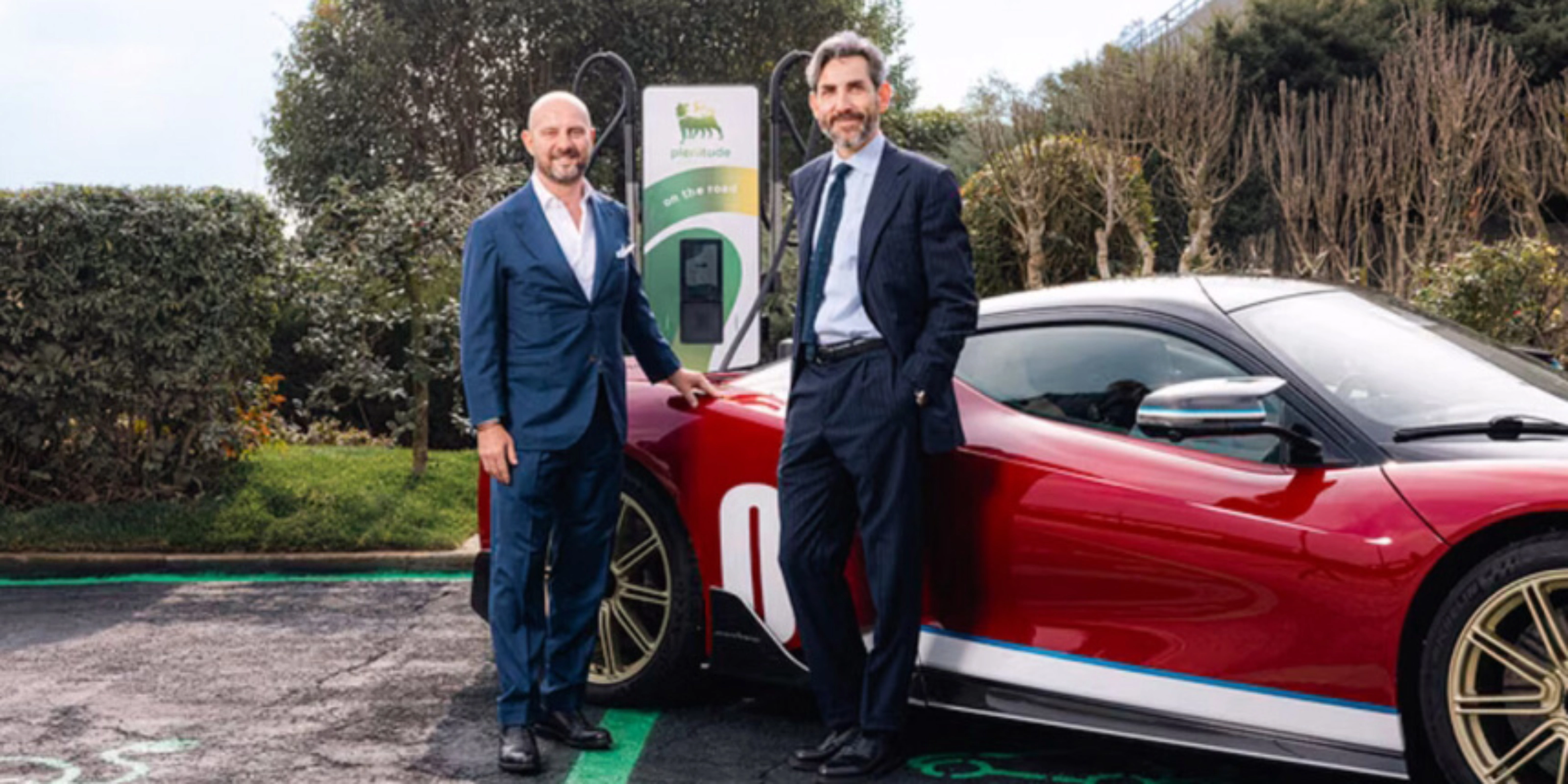 Two men in suits stand beside a red electric sports car at a charging station.