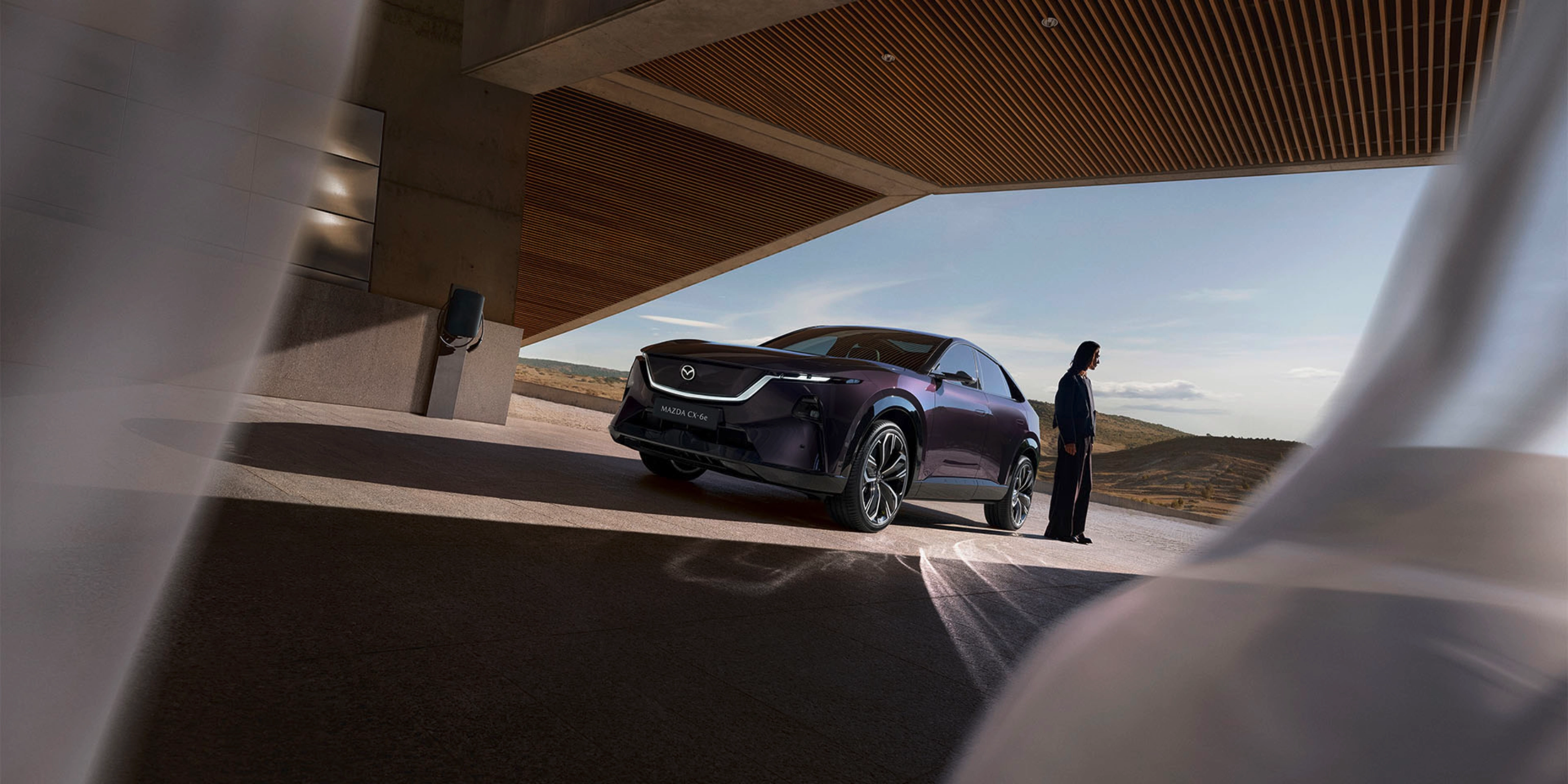 A person stands near a dark Mazda SUV under a modern building with open landscape behind.