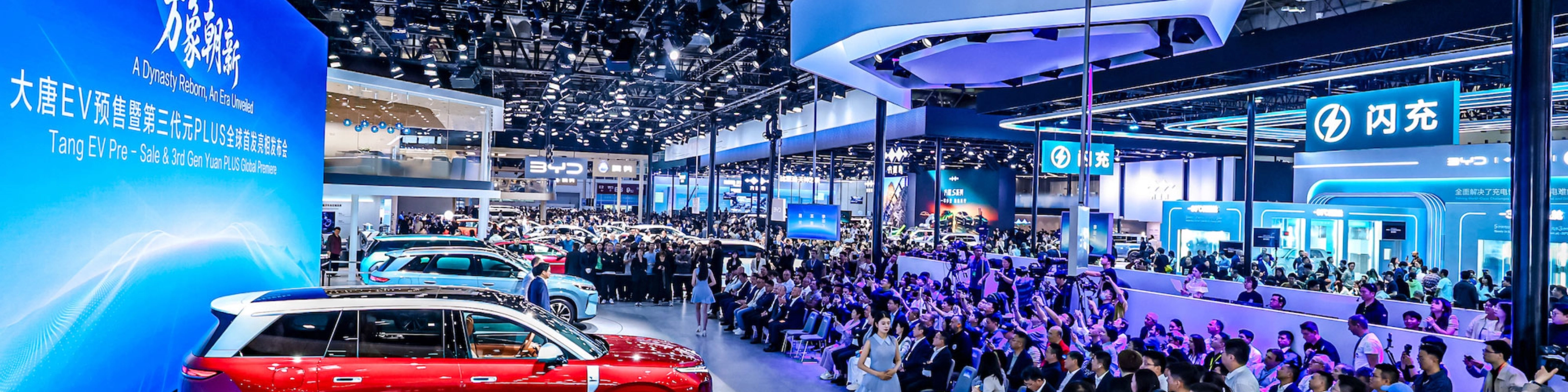 Red car on display at a crowded auto show with large screen.