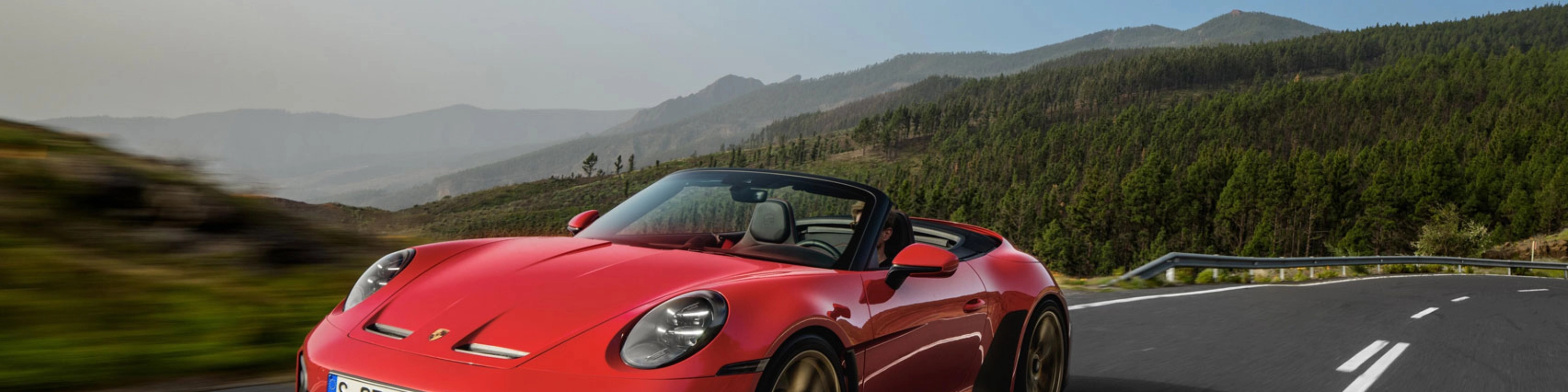 Red Porsche convertible driving on a winding road through green hills.