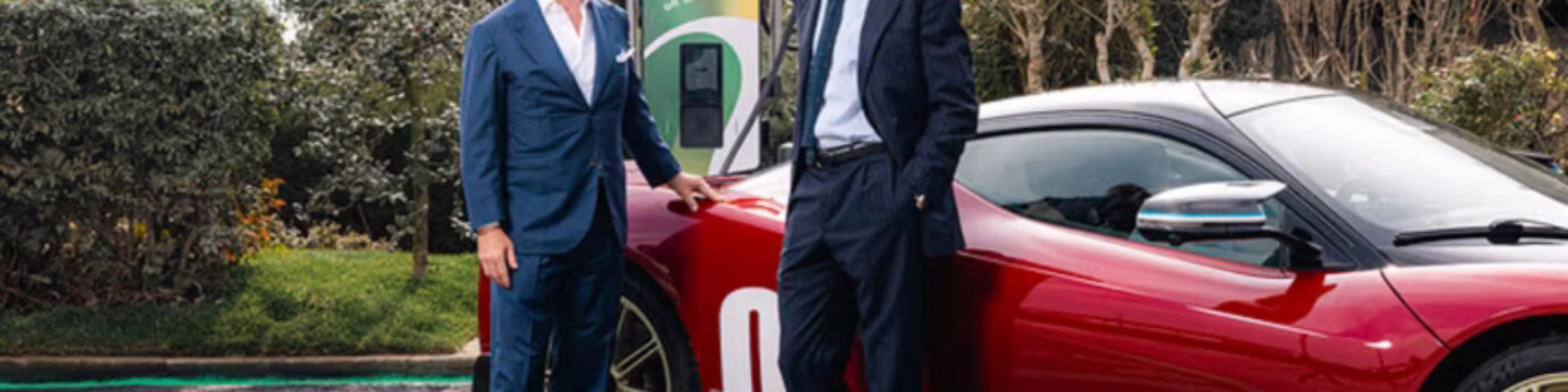Two men in suits stand beside a red electric sports car at a charging station.