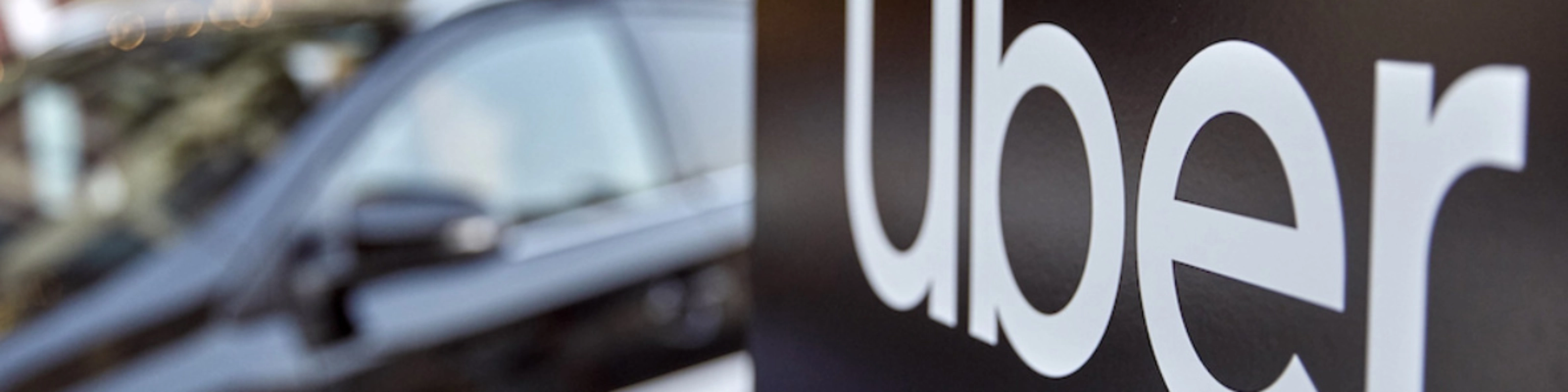 Uber-Logo auf einem Schild, dahinter unscharf ein Auto.