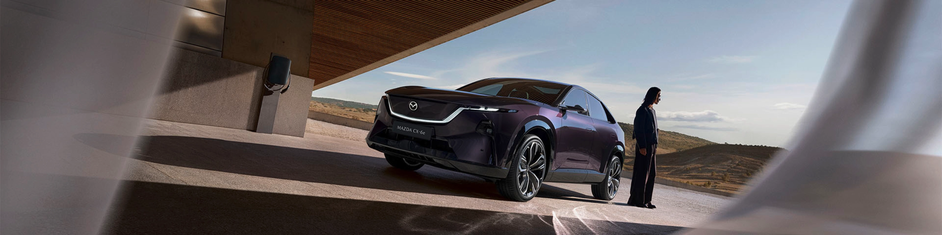 A person stands near a dark Mazda SUV under a modern building with open landscape behind.