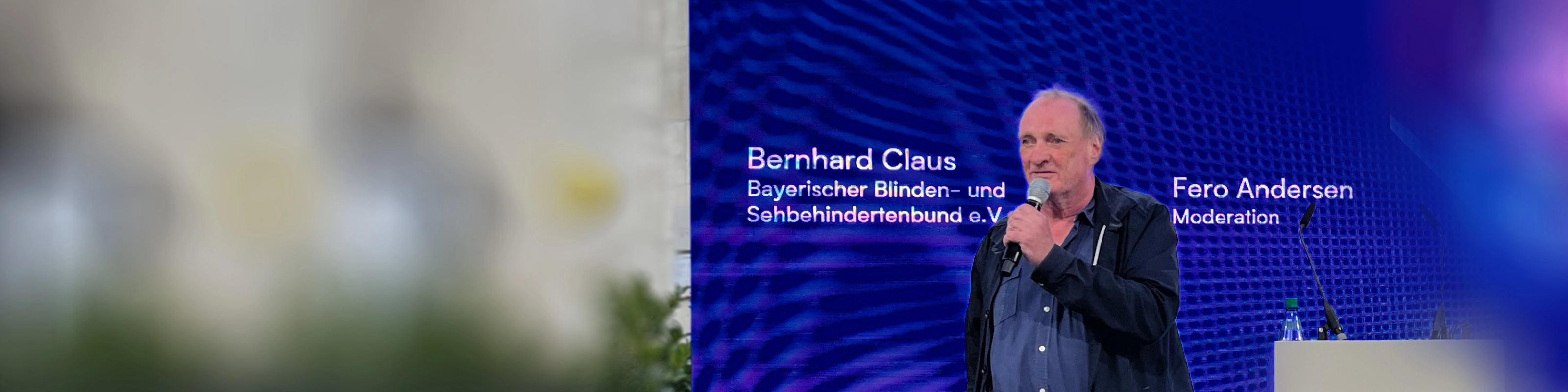 Ein Redner steht auf einer Bühne vor einem Bildschirm zur Mobilität für sehbehinderte Menschen.