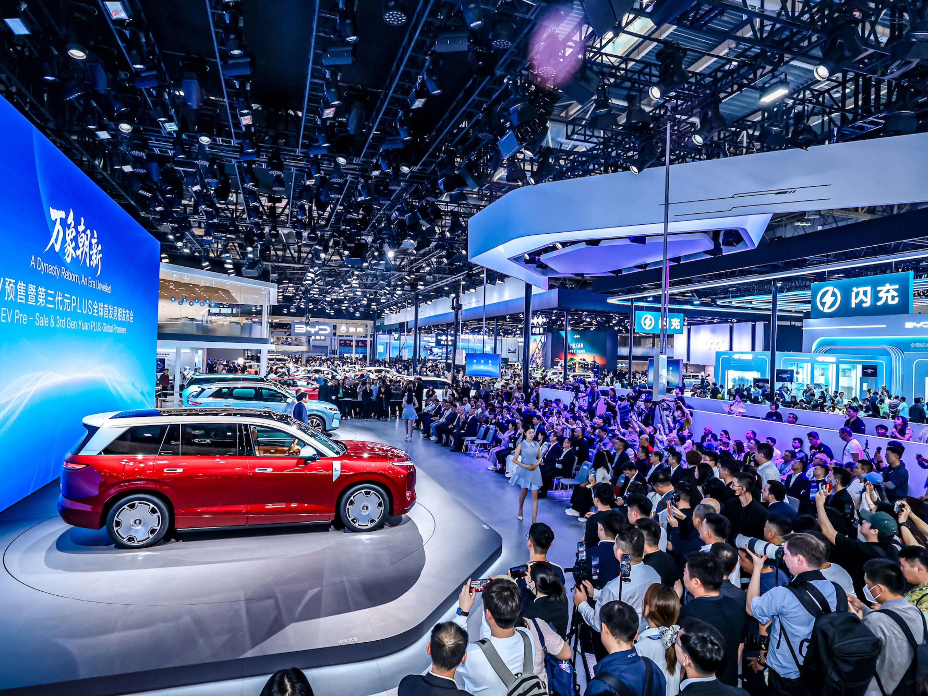 Red car on display at a crowded auto show with large screen.