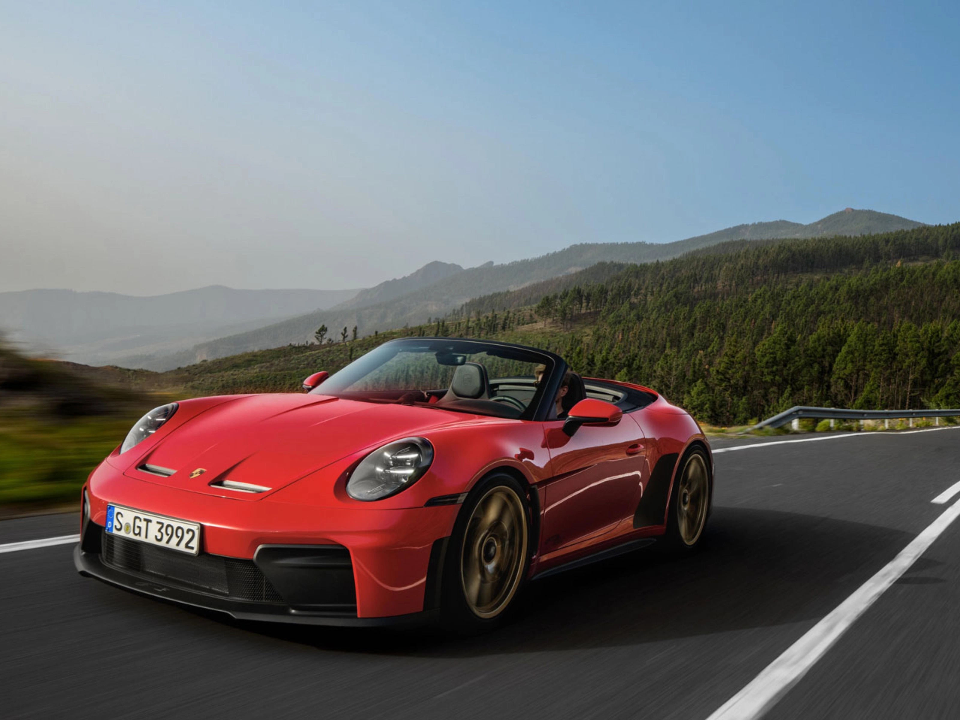 Red Porsche convertible driving on a winding road through green hills.