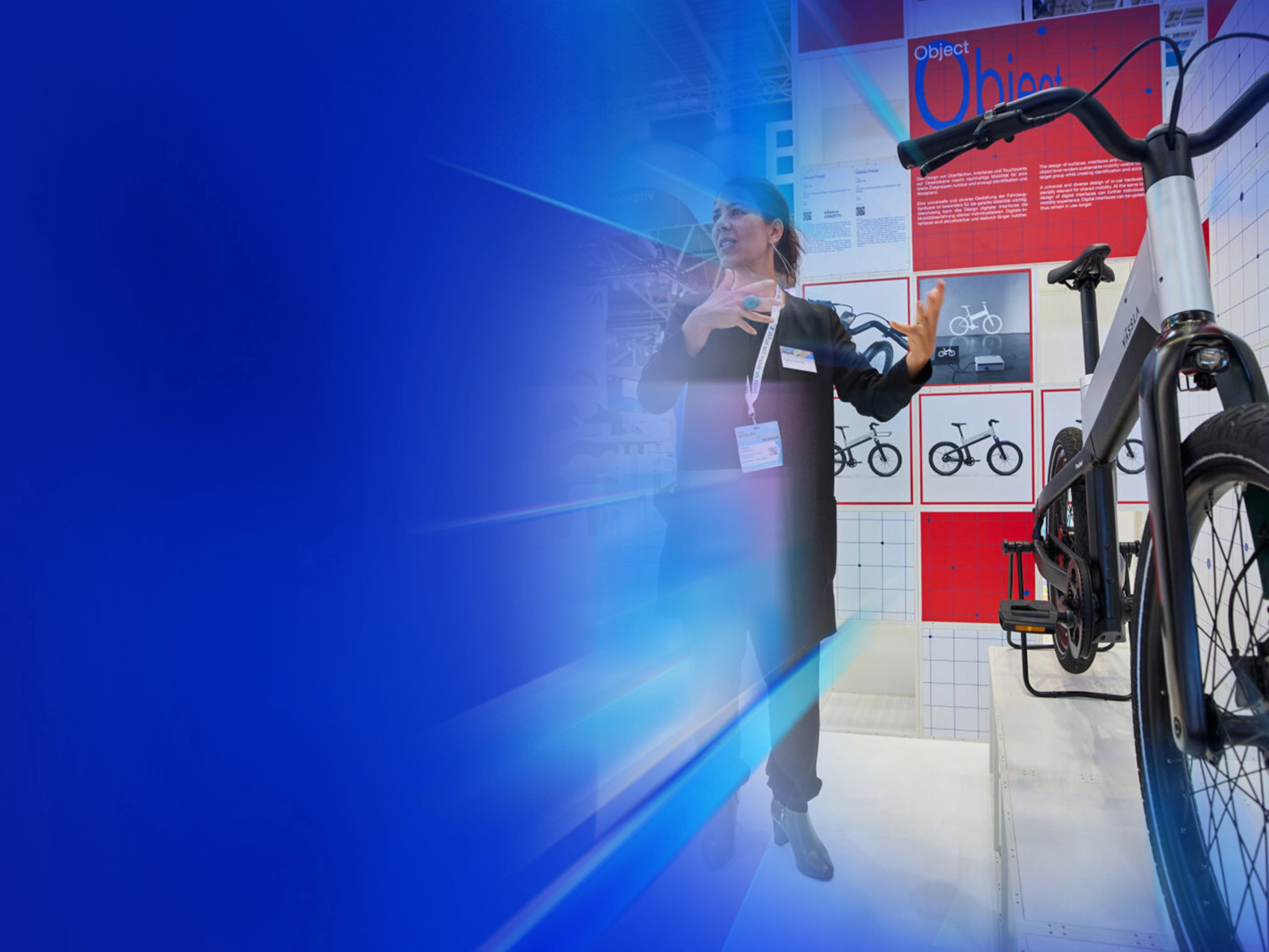 Eine Frau präsentiert ein modernes Fahrrad an einem Messestand mit Designpostern im Hintergrund.