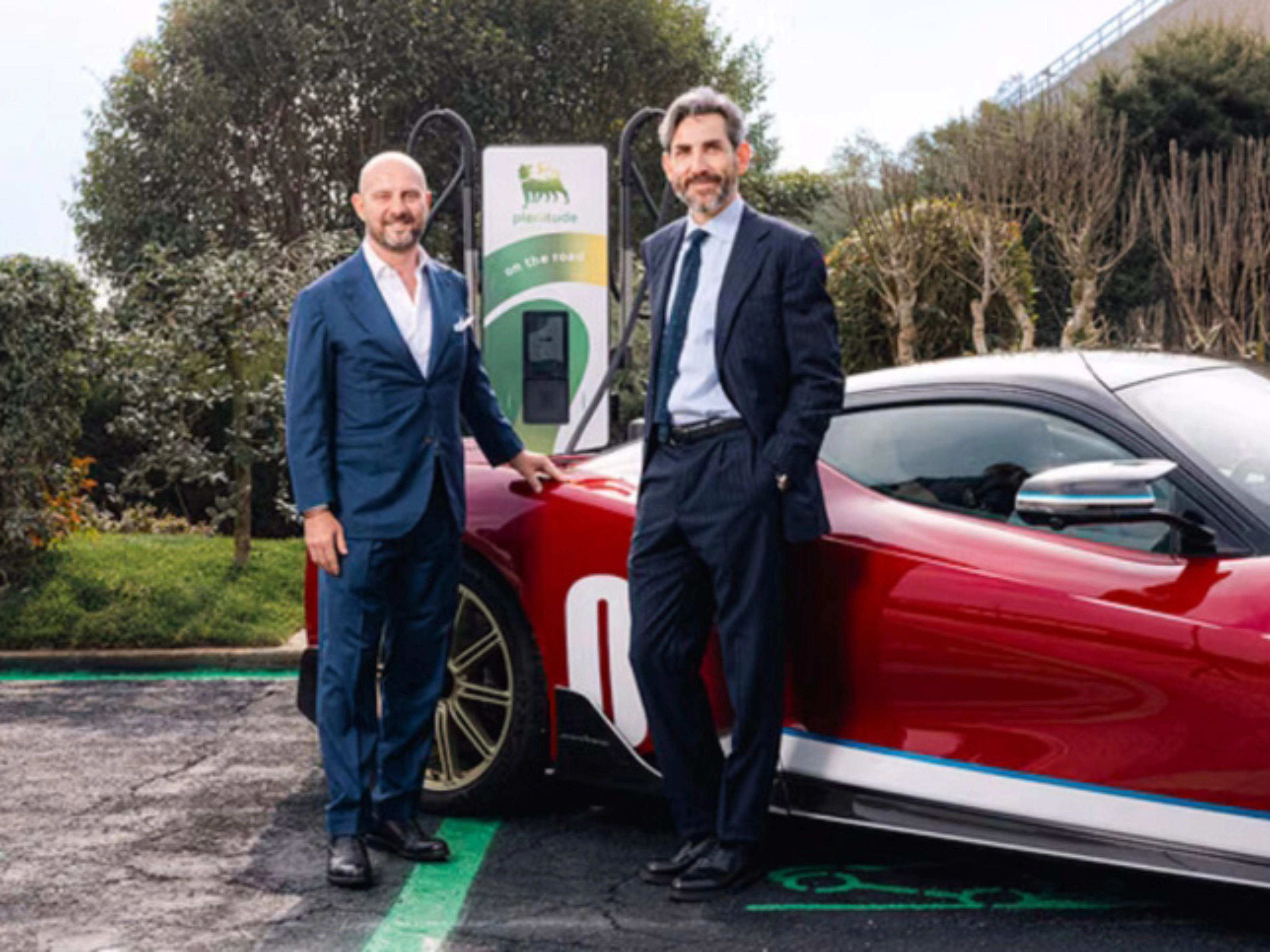 Two men in suits stand beside a red electric sports car at a charging station.