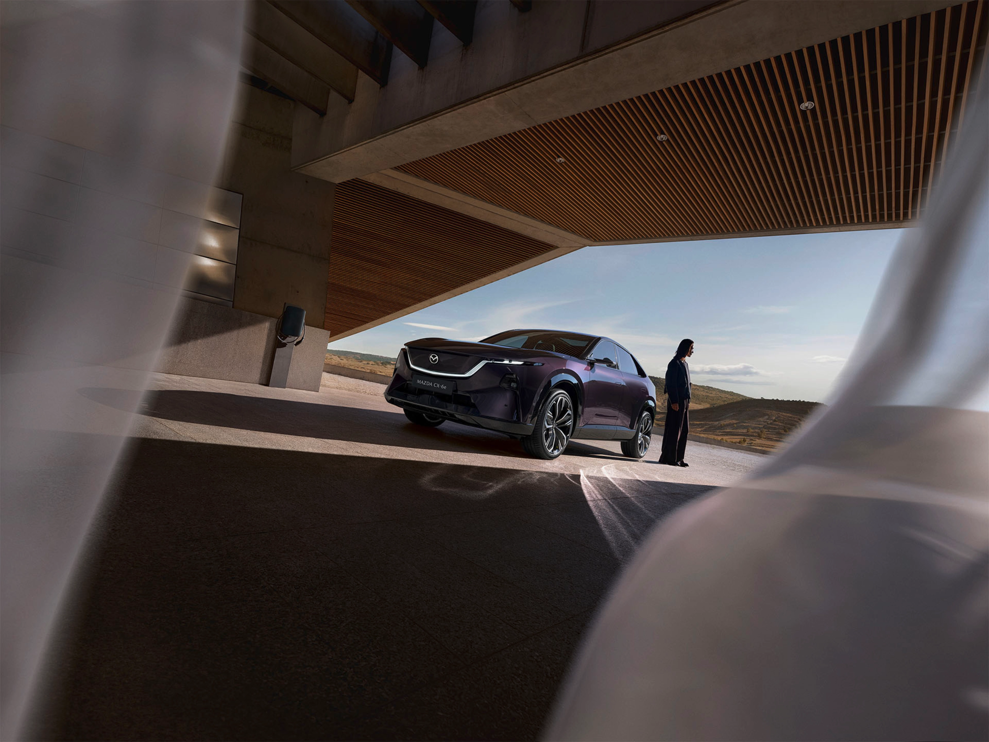 A person stands near a dark Mazda SUV under a modern building with open landscape behind.