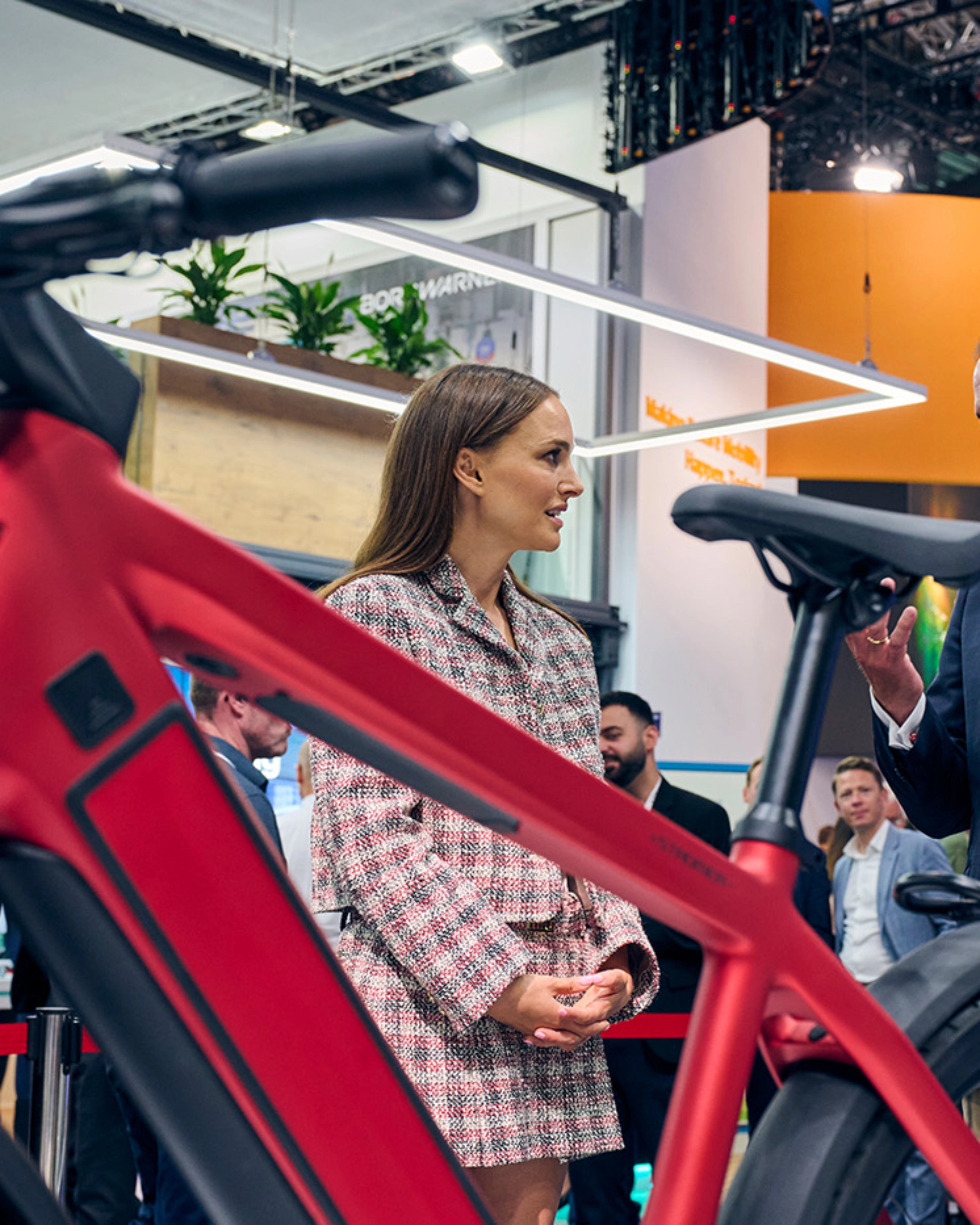 Natalie Portman in conversation with exhibitors at IAA Mobility 2023, standing near a red bicycle in a trade fair setting.