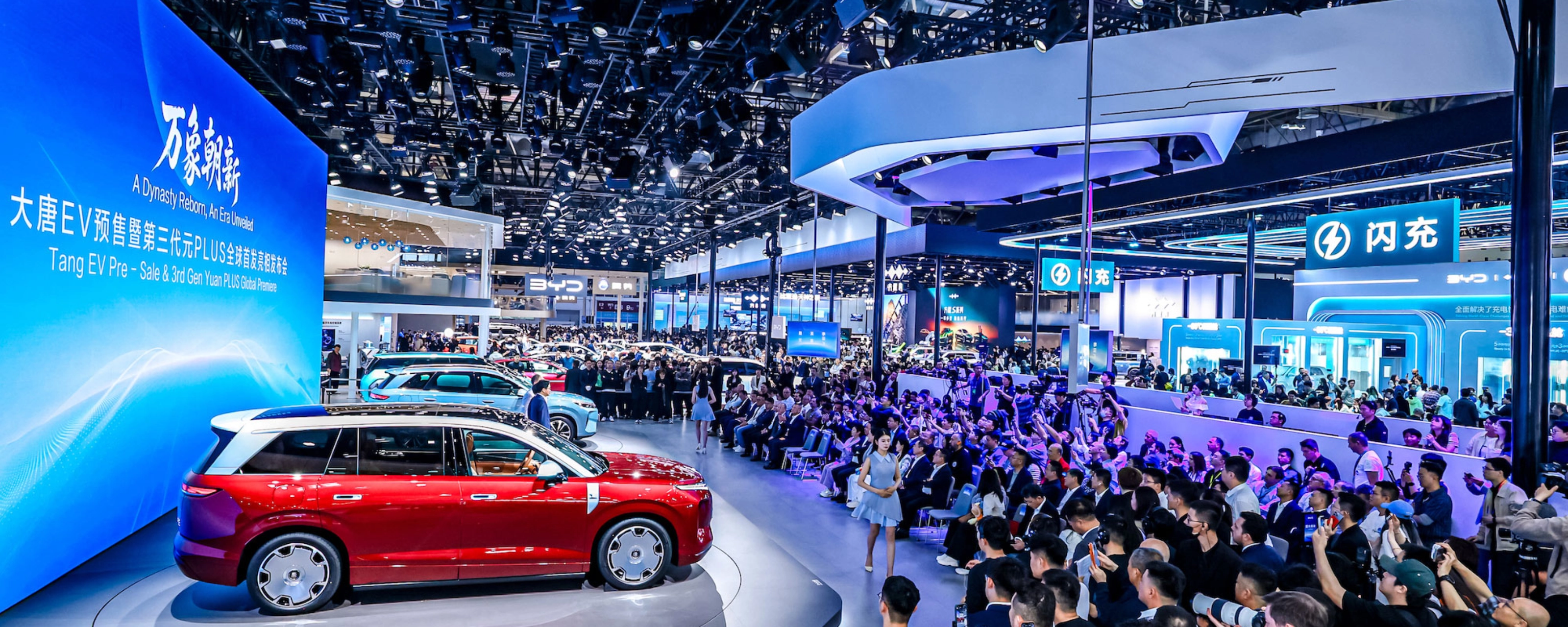 Red car on display at a crowded auto show with large screen.