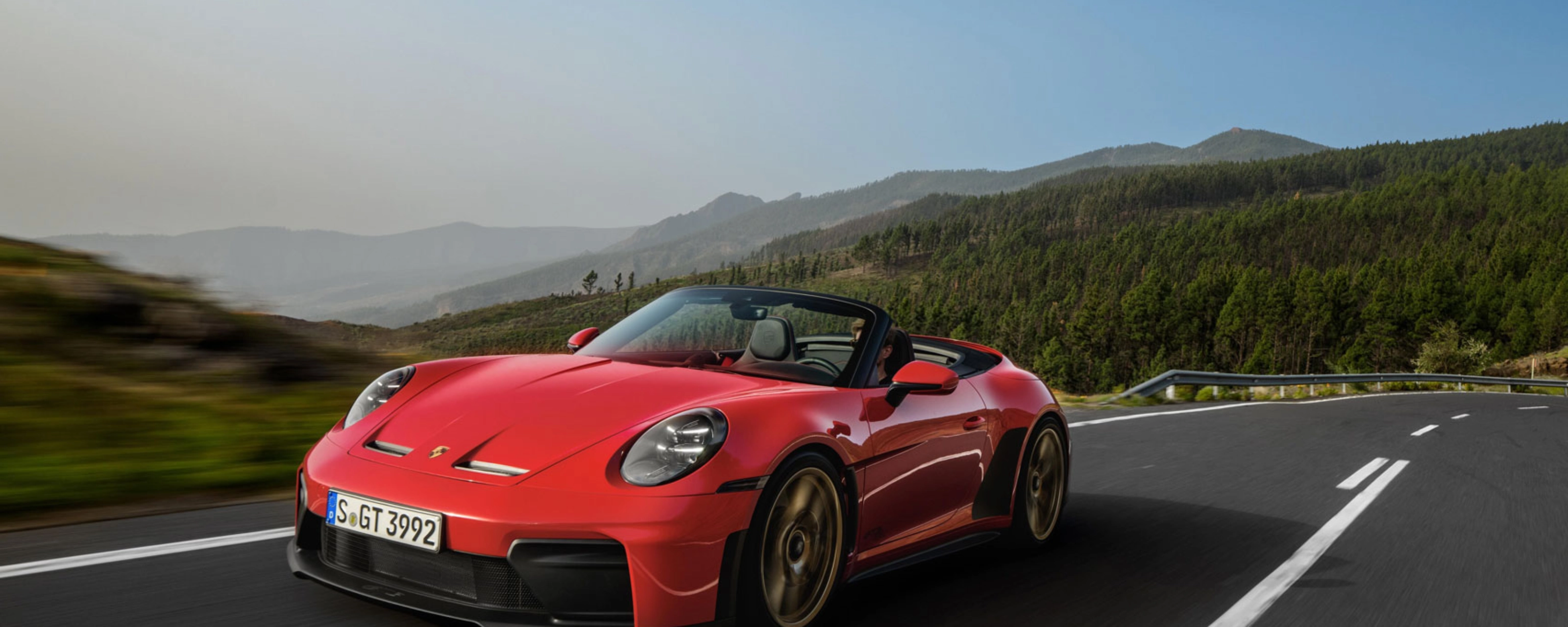 Red Porsche convertible driving on a winding road through green hills.