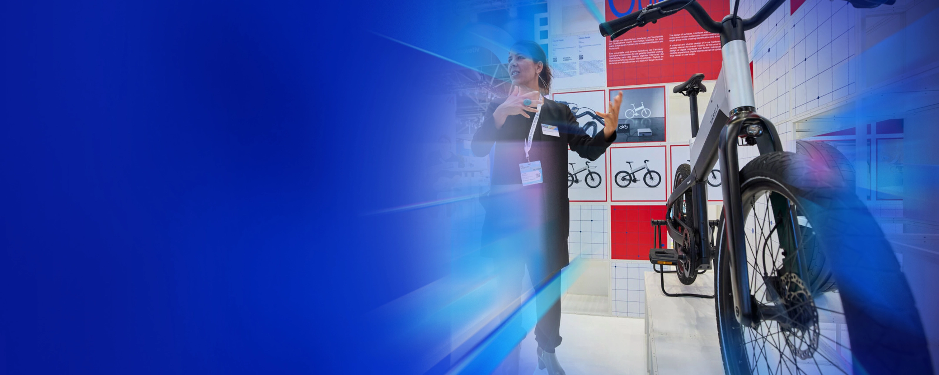 Eine Frau präsentiert ein modernes Fahrrad an einem Messestand mit Designpostern im Hintergrund.