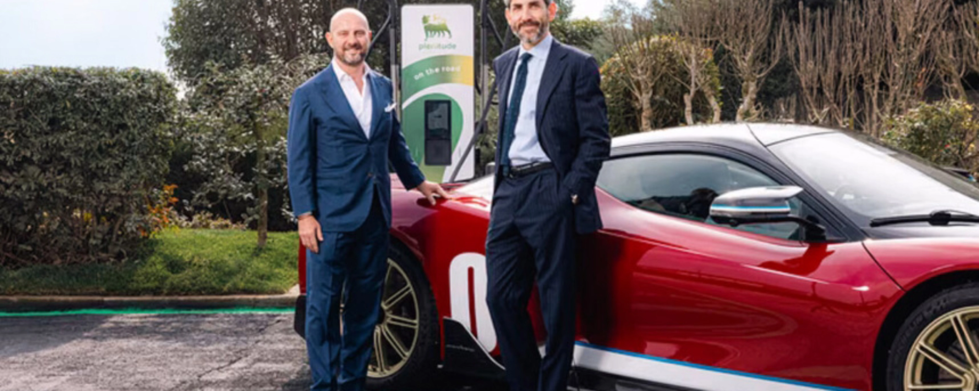 Two men in suits stand beside a red electric sports car at a charging station.