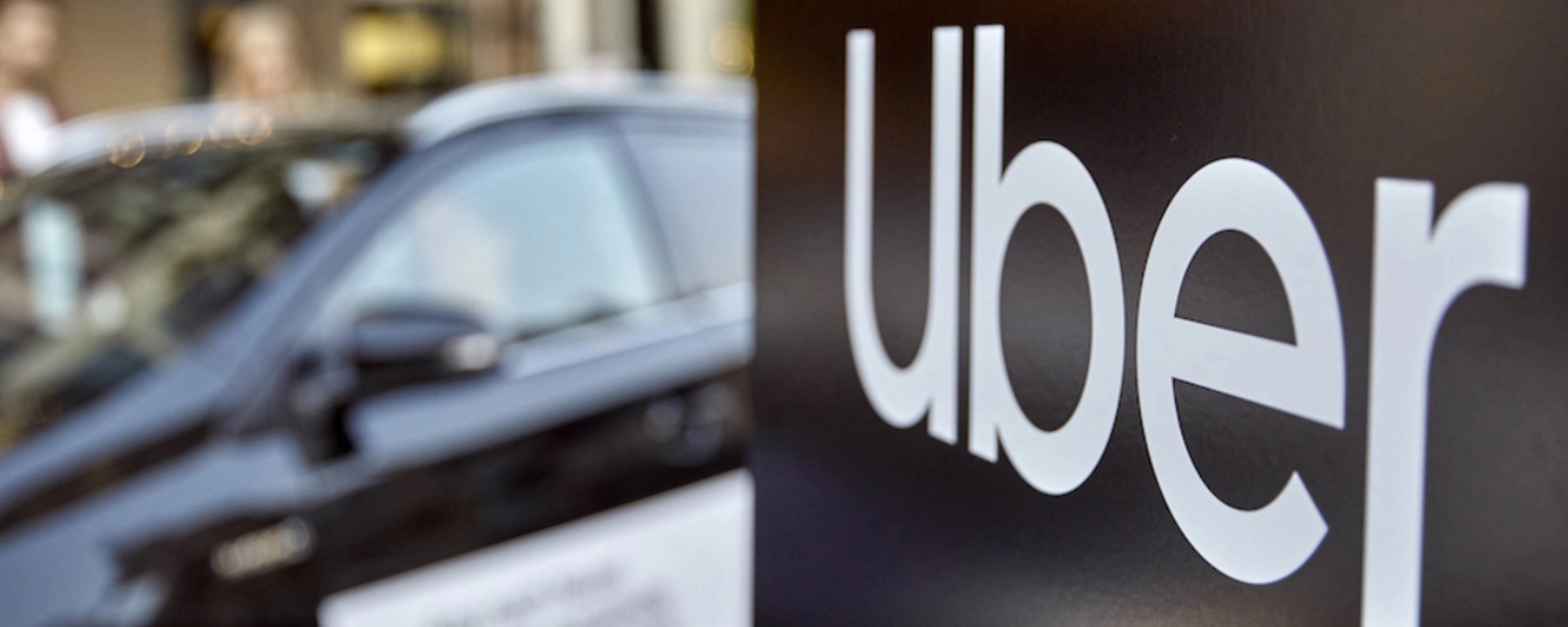 Uber-Logo auf einem Schild, dahinter unscharf ein Auto.