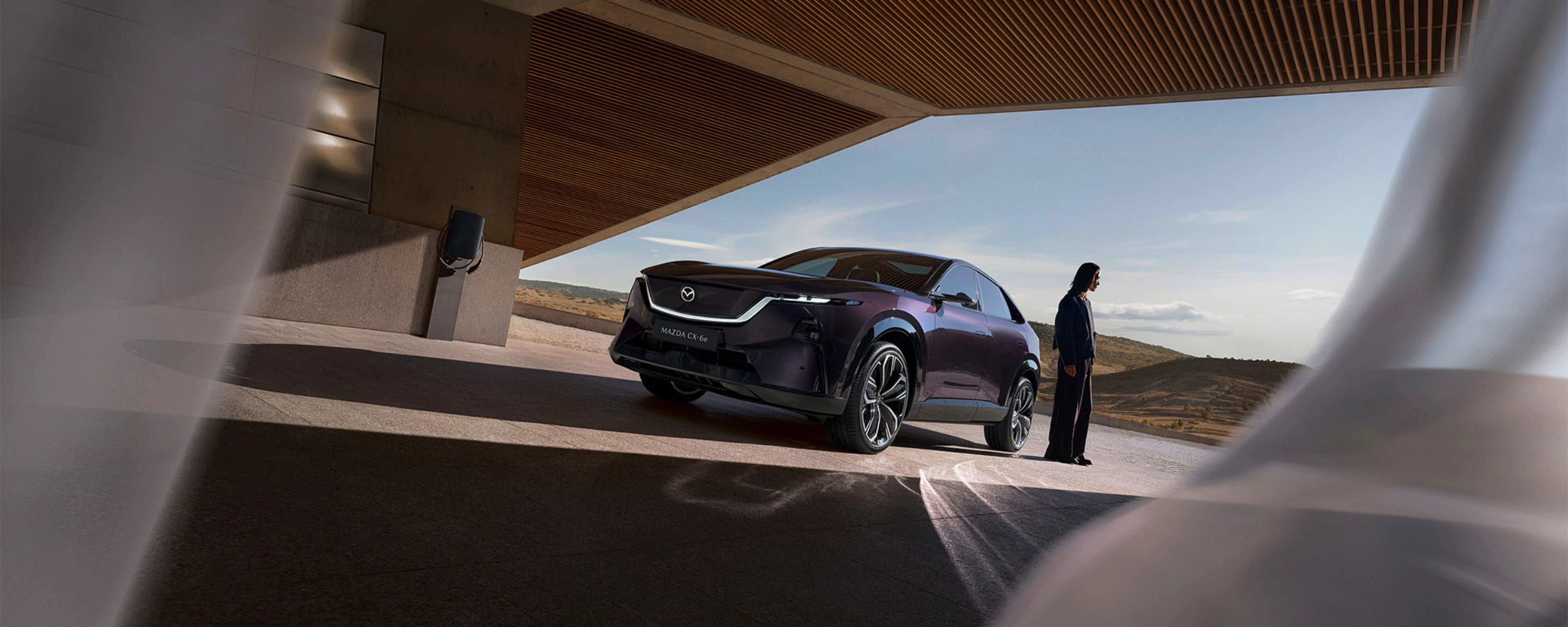 A person stands near a dark Mazda SUV under a modern building with open landscape behind.