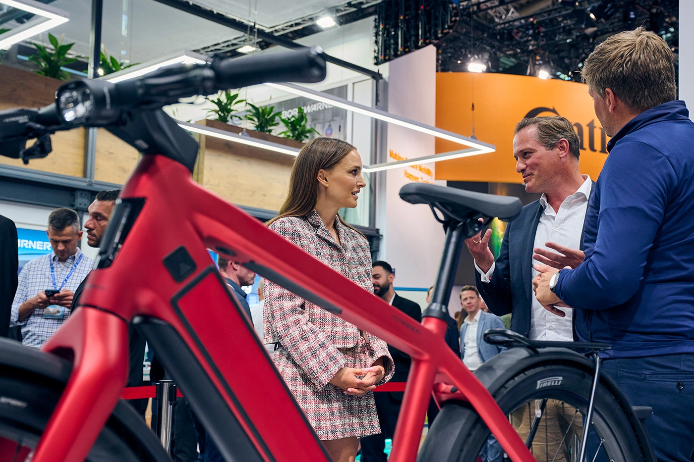 Natalie Portman in conversation with exhibitors at IAA Mobility 2023, standing near a red bicycle in a trade fair setting.