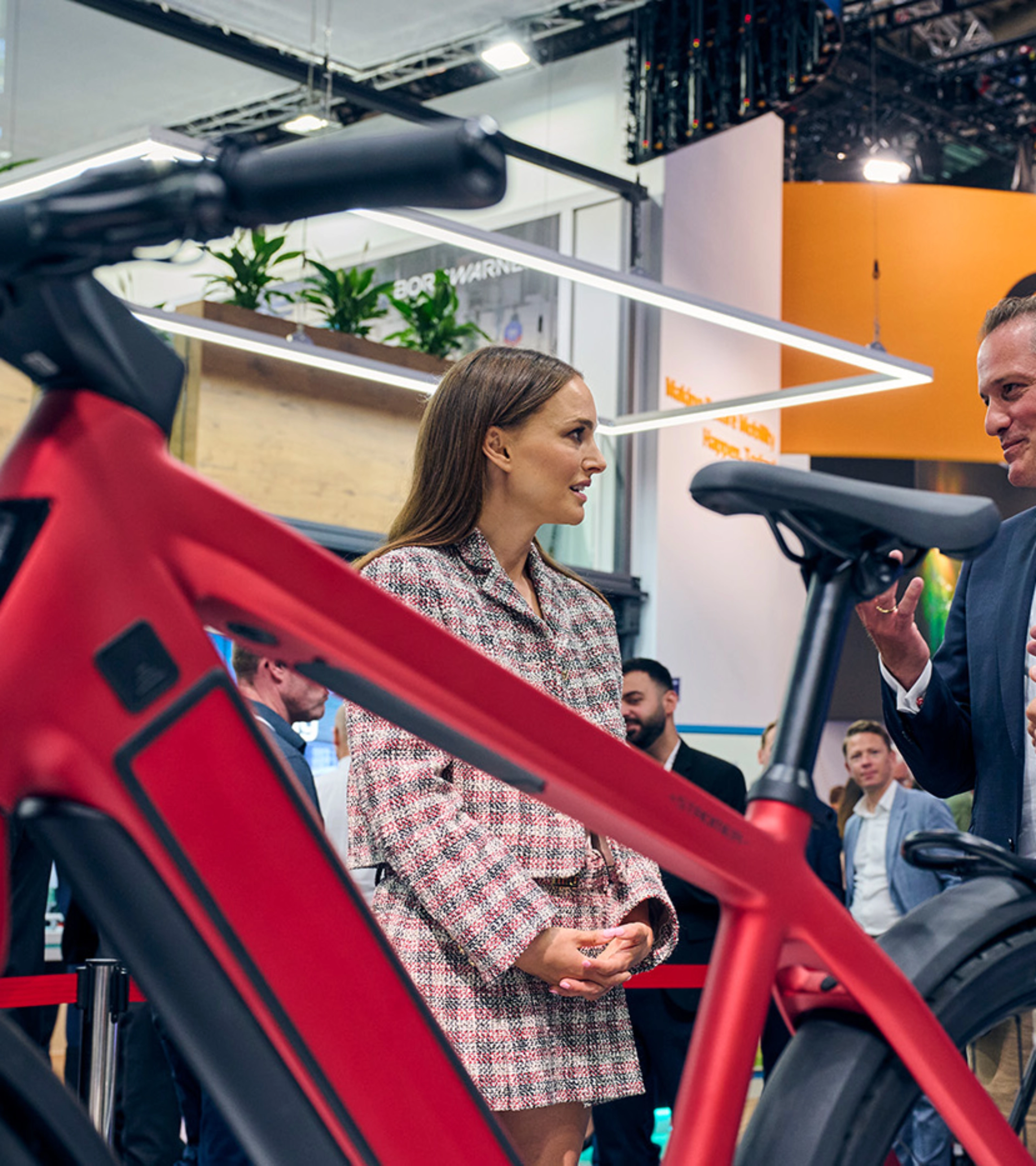Natalie Portman in conversation with exhibitors at IAA Mobility 2023, standing near a red bicycle in a trade fair setting.