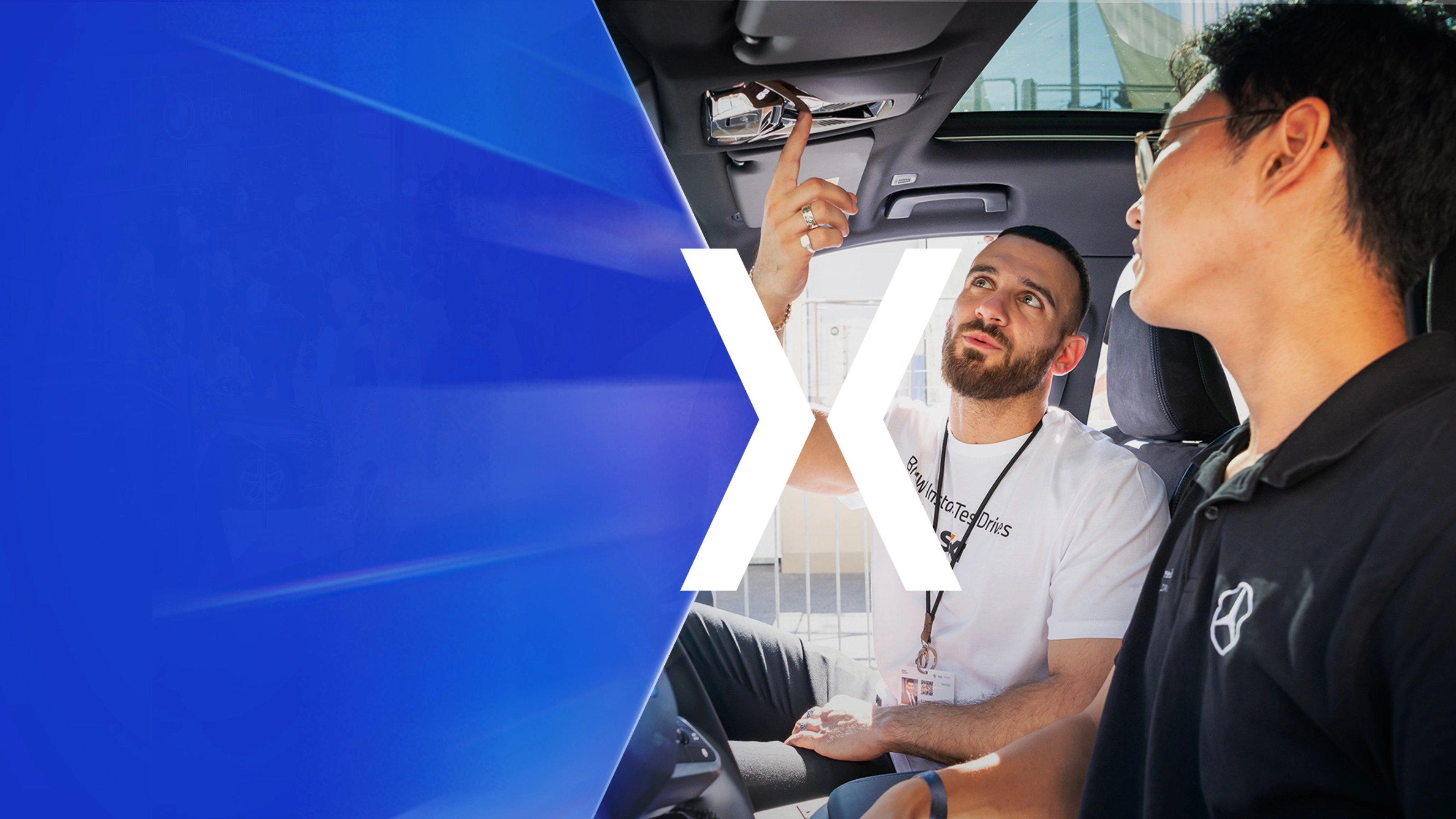 Two men sit in a car while one of them explains a control on the ceiling panel.