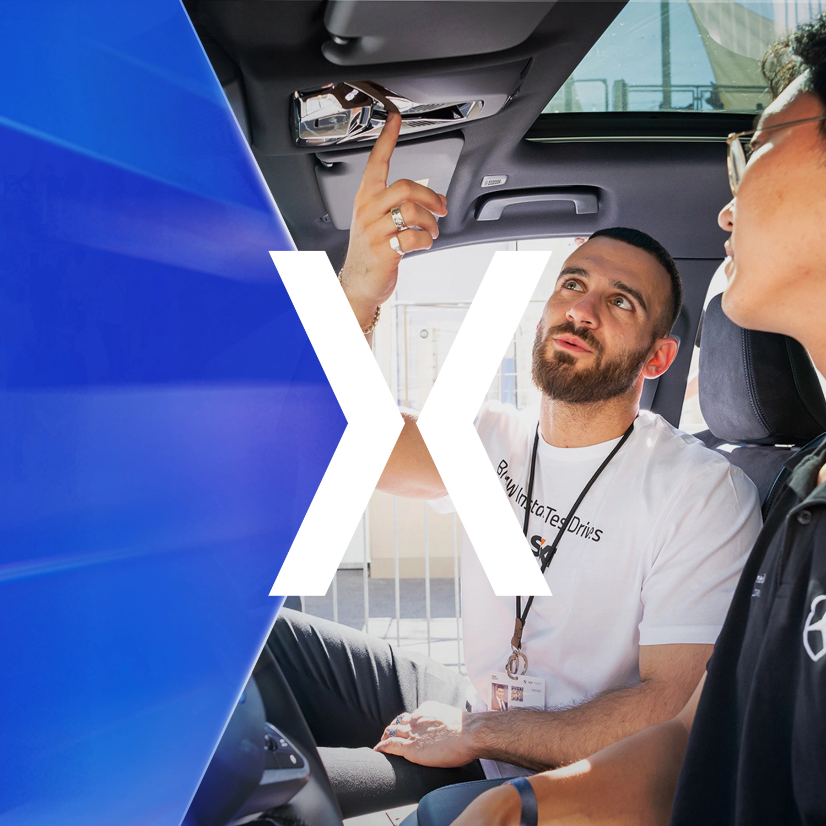 Two men sit in a car while one of them explains a control on the ceiling panel.
