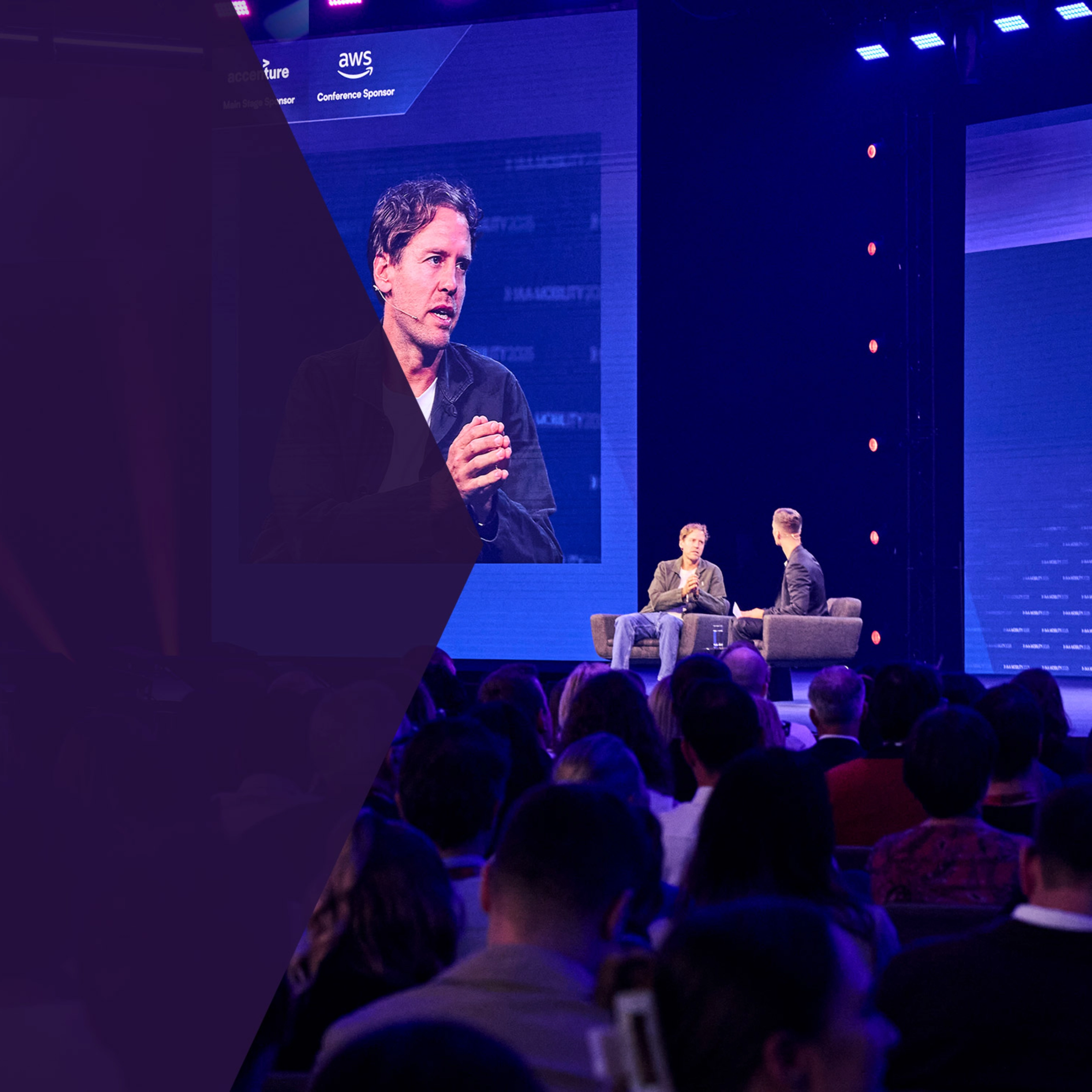 Two men talk on stage during a conference, with one shown on a large screen behind them.