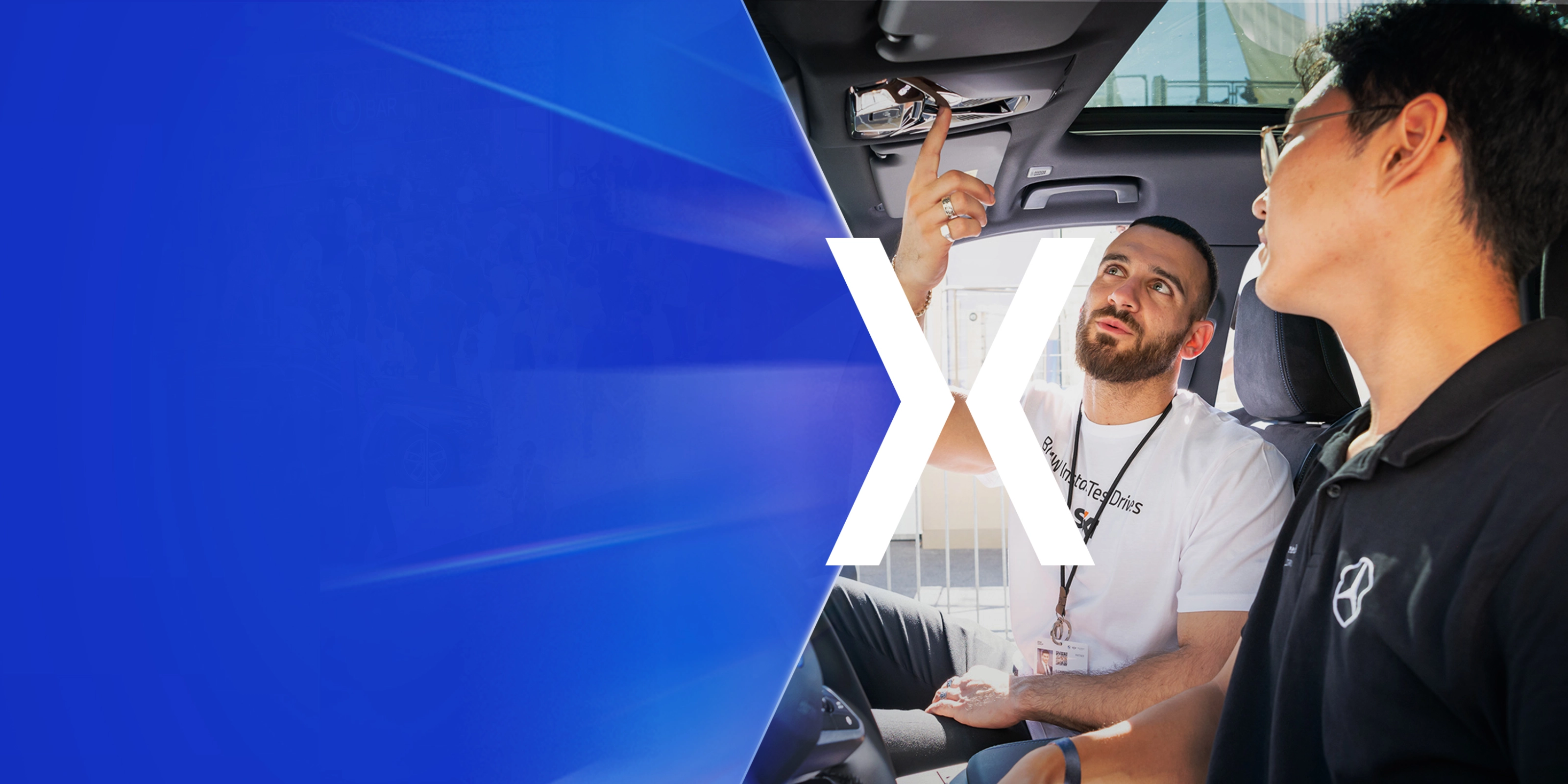 Two men sit in a car while one of them explains a control on the ceiling panel.